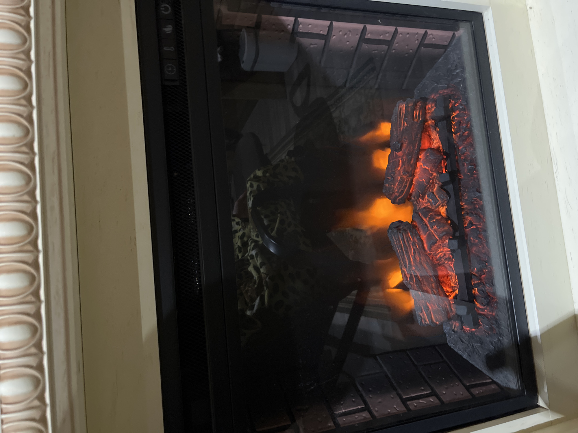 The image shows an electric fireplace with realistic-looking logs and flames. The fireplace is housed in a cream-colored mantelpiece with decorative molding. The interior of the fireplace features a brick-patterned backing and a glass front that reflects objects in the room, including a leopard-print fabric and a small white cylindrical object. The logs themselves are dark and appear to be glowing red and orange, simulating embers. There are also visible controls on the top right of the fireplace, including what appears to be a power button and other function icons. The scene is indoors, and the lighting suggests it may be evening or a dimly lit room. There is no information in the image that could indicate the location is in Saratov, Russia.
