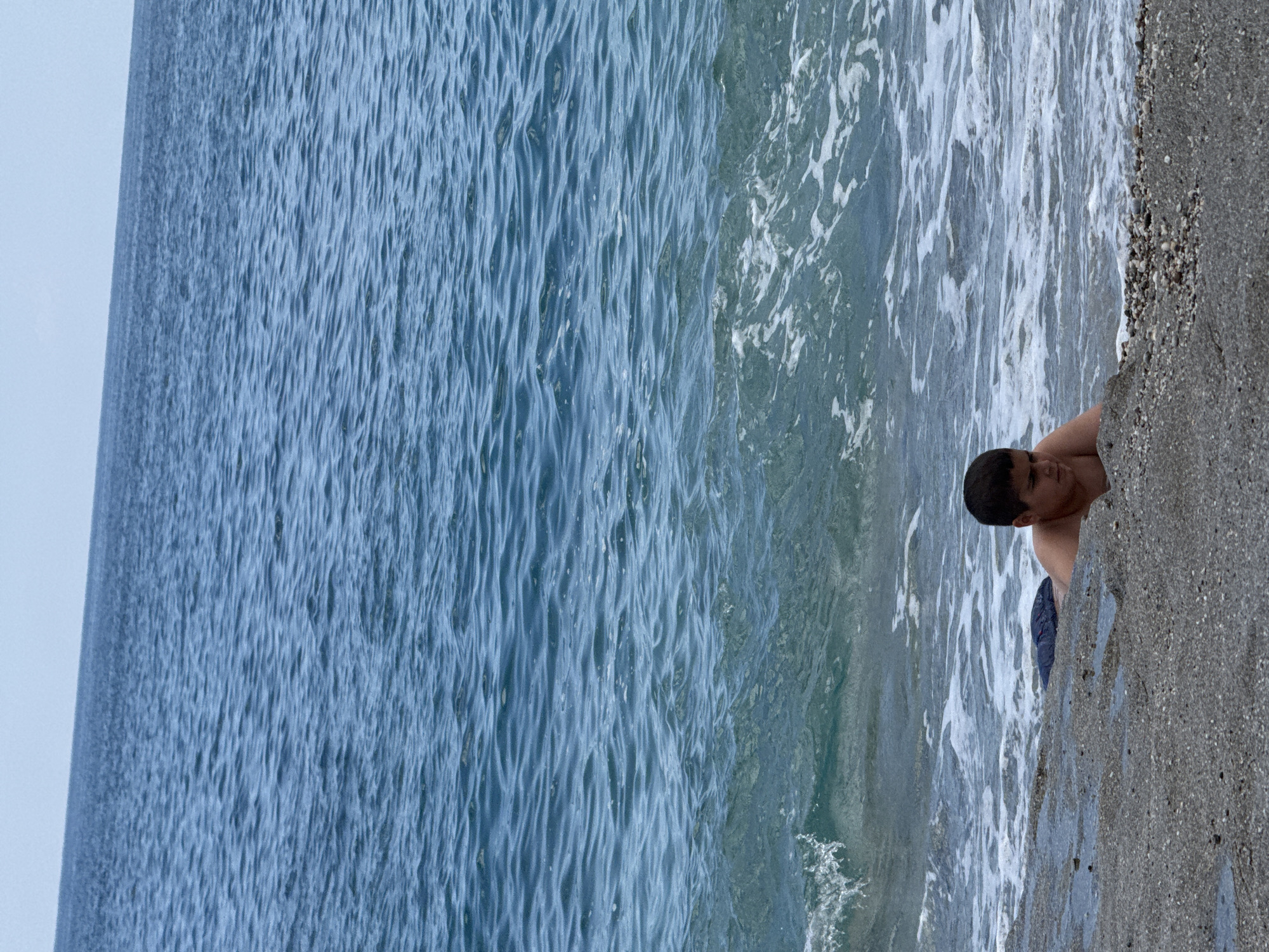 A vertical outdoor shot depicts a beach scene. A young boy with dark hair and tanned skin is partially submerged in the water at the edge of the beach. He is wearing blue shorts. The water is a mix of blue and white, with waves breaking near the shore. The beach itself is a mix of sand and small rocks. The location and city are unknown.