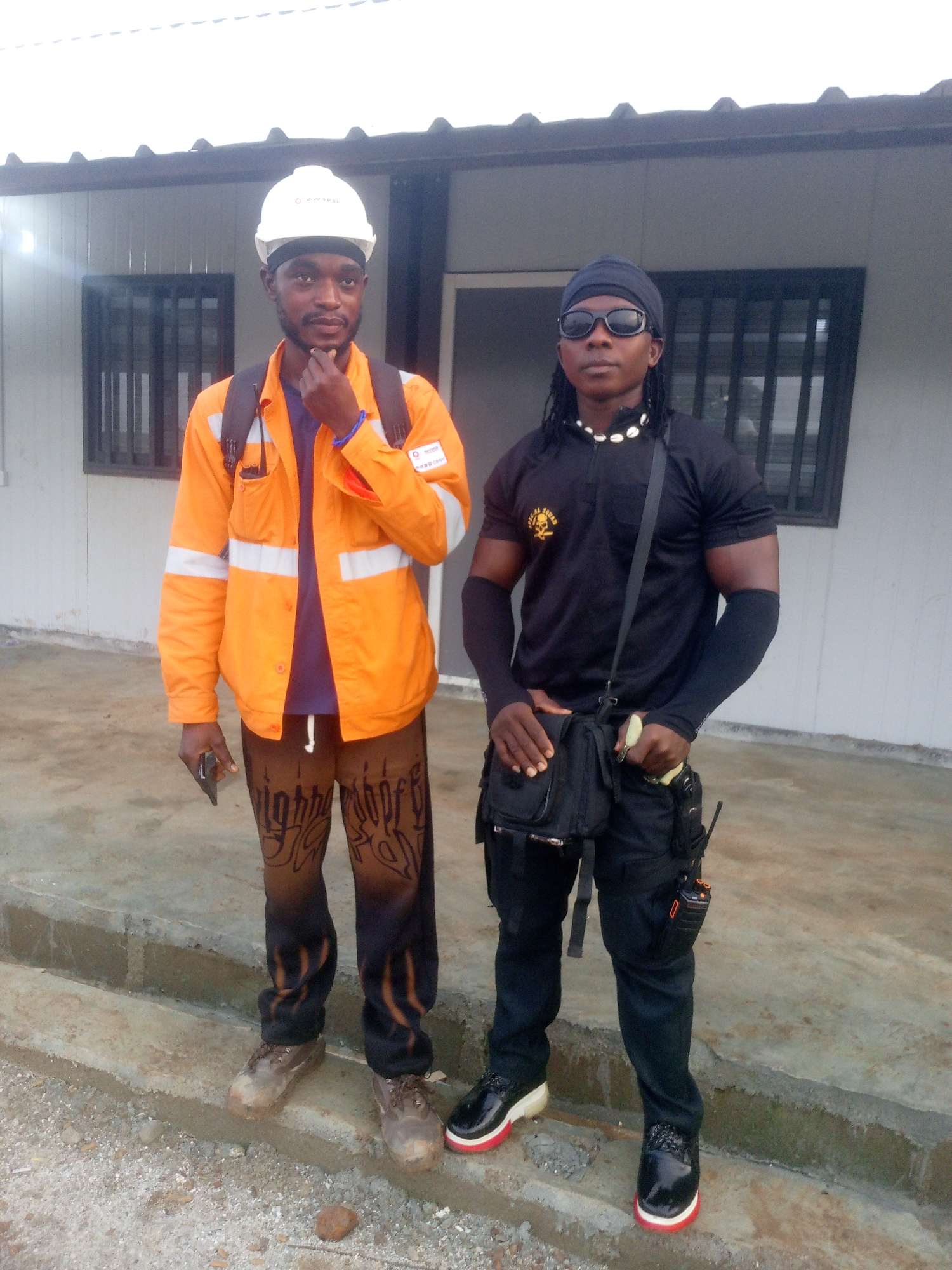 Two men stand in front of a prefabricated building. The man on the left wears a white hard hat, an orange high-visibility jacket over a purple t-shirt, and patterned brown pants. He holds a dark object, possibly a mobile phone, in his left hand. The man on the right wears a black durag, sunglasses, a black t-shirt with a yellow logo, black arm sleeves, black pants, and black shoes with red soles. He has a cross-body bag and a utility belt with several pouches and a walkie-talkie. The ground in front of them is concrete and gravel. The building appears to be a modular structure, possibly for a construction site or temporary facility, with visible windows and a corrugated roofline. The lighting suggests it is daytime. The overall impression is that of a worksite or industrial area.