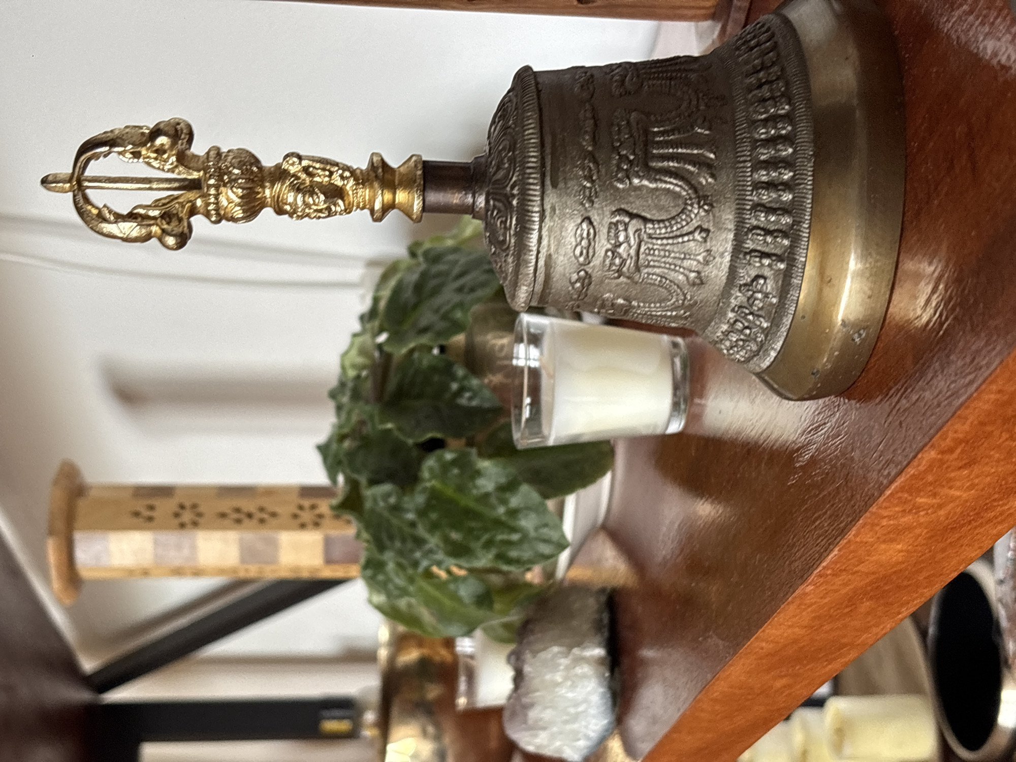 The image displays a close-up, static shot of a decorative arrangement on a wooden shelf, presumably indoors in Mexico City, Mexico.
The central and most prominent subject is a Tibetan ritual bell (ghanta) with an ornate vajra (dorje) handle. The handle, appearing golden or brass-colored, features intricate details including a five-pronged vajra at the top and a small, stylized figure in the middle, likely a deity or Buddha, surrounded by further decorative elements. The bell itself is a darker, metallic gray or antique bronze color, heavily embossed with elaborate designs that include what appear to be dragons or mythical creatures, and auspicious symbols encircling its body. The bell rests on a polished, reddish-brown wooden shelf, which shows a reflective sheen.
Behind the bell, to its left, is a small potted plant with dark green, somewhat crinkly leaves, partially obscuring a blurred, lighter-colored object. To the right of the bell, a small, clear glass votive holder contains a white, unlit candle. Further to the right, a translucent, rough-textured object, likely a raw crystal or mineral specimen, is visible.
In the blurred background to the far left, a tall, narrow wooden object with a checkered or patterned design is partially visible. The overall background is a plain, light-colored wall. Below the main shelf, other out-of-focus items, including dark rectangular shapes and another light-colored object, are faintly discernible. The lighting appears soft and even, suggesting an indoor setting with no specific time of day indicated. No discernible text is visible on any of the objects. The scene conveys a sense of curated decoration, possibly of a spiritual or aesthetic nature.