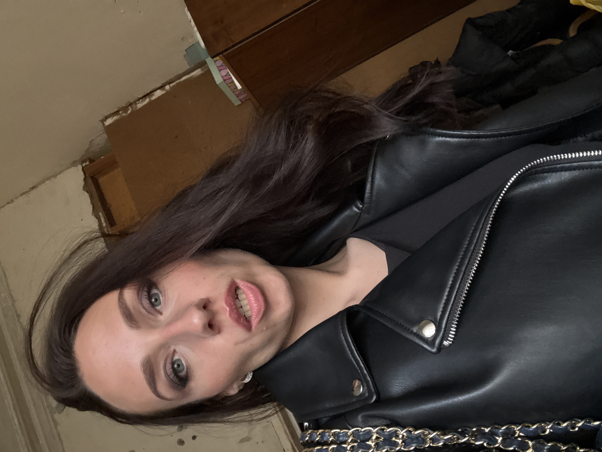 A woman with long brown hair, light skin, and green eyes is pictured wearing a black leather jacket with silver hardware and a black top. The environment appears to be an indoor space with deteriorated walls and woodwork. It is impossible to determine the location in Saint Petersburg, Russia from the image.
