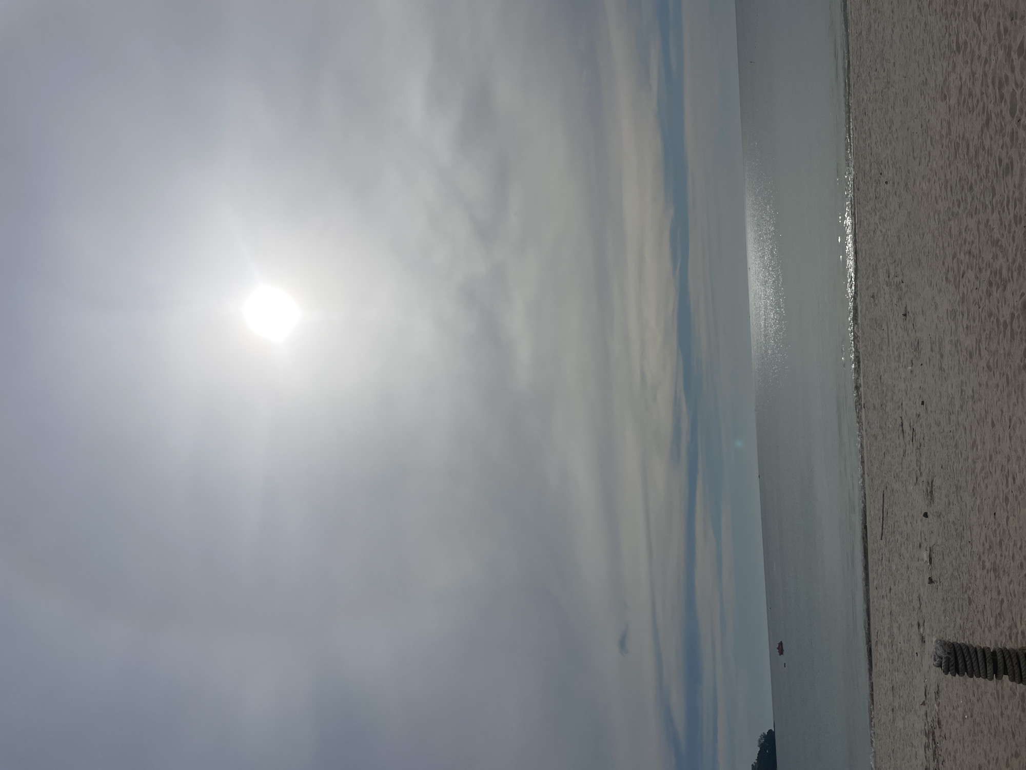 The image depicts a bright, sunny day at Bang Tao Beach in Choeng Thale, Thailand. The sky is mostly covered with clouds, with the sun shining through. The beach is sandy, and the water is calm. A small boat is seen in the distance. A rope is visible in the lower-right corner, on the sand.