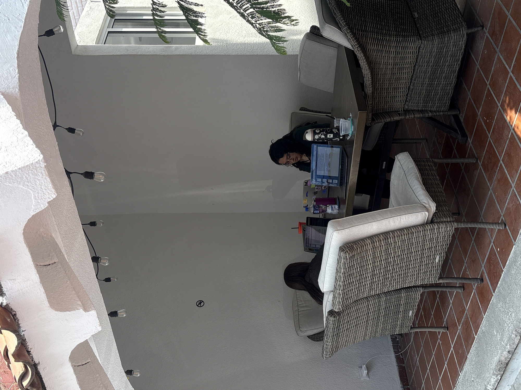 The image captures a shaded outdoor patio or terrace, oriented 90 degrees clockwise, where two individuals are seated at a table, working on laptops during the daytime. Correctly oriented, a white, textured ceiling and wall structure, adorned with hanging string lights, would be on the left, leading to a terracotta-tiled floor on the right.
One woman with dark hair and glasses is clearly visible, leaning forward and focused on her laptop screen. Next to her laptop on the dark-colored table are a water bottle or thermos with white illustrations and a clear cup. The second person is partially seen from the back, also seated at the table in a wicker chair with light-colored cushions, seemingly engaged with another laptop. The chairs are made of brown woven wicker.
Natural light enters the space, primarily from a window in the background where green, pine-like foliage is visible. The scene suggests a calm environment for work or study. A small, indistinct black circular symbol with a line through it is present on a white wall in the mid-ground. A white electrical outlet is visible near the floor. No distinct text is readable on any objects or screens. The setting is consistent with a pleasant outdoor area, potentially in Puebla, Mexico.