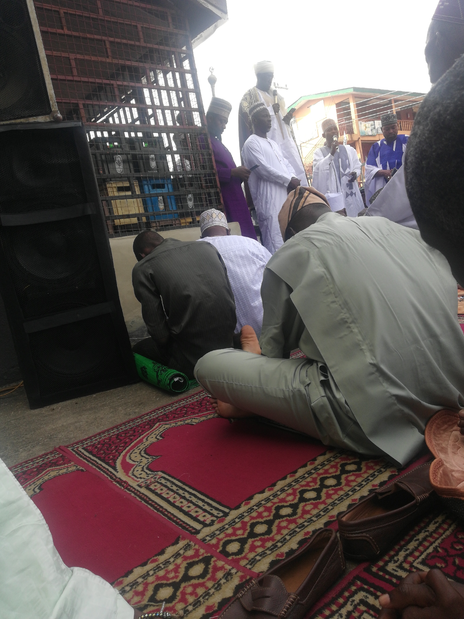 The image shows a group of Muslim men gathered on prayer mats, presumably praying. Some of the men are kneeling or sitting on the mats, while others are standing and appear to be leading the prayer or giving a sermon. Several men are wearing traditional Muslim attire, including white robes and hats. A large speaker is visible on the left side of the image. Behind the praying men, there are some metal grates, and what looks like crates stacked together. The location appears to be an outdoor space in an urban area, based on the surrounding buildings.