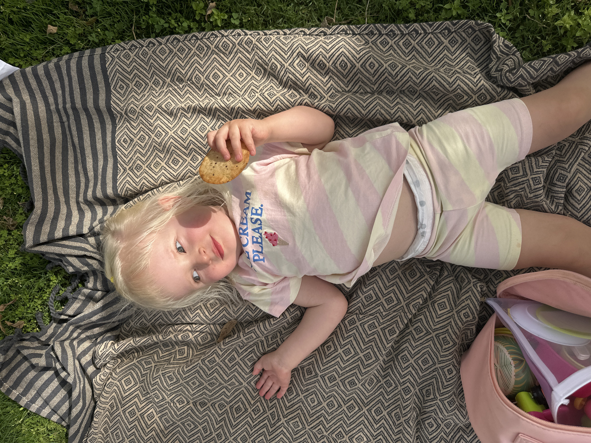 A young child with blonde hair lies on a patterned blanket on grass. The child is wearing a pink and yellow striped shirt that reads "ICE CREAM PLEASE." They are holding a round cracker in their right hand. A pink bag sits beside them on the blanket. The setting is outdoors, with grass visible around the blanket. There is no evidence of Petah Tikva, Israel in the image.