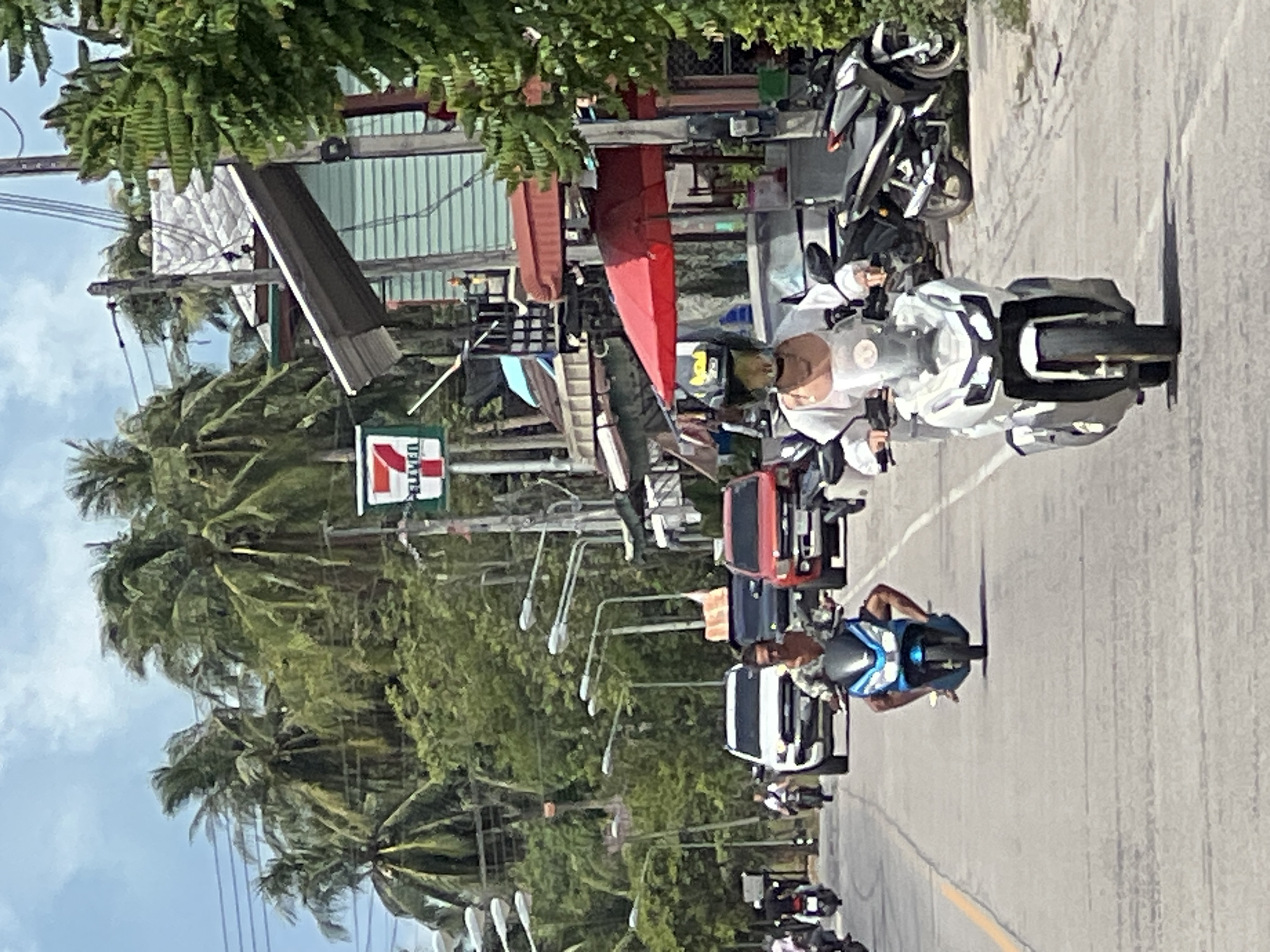 The image shows a street scene in Tambon Ko Pha Ngan, Thailand, on a partly cloudy day. In the foreground, a person wearing a helmet and a white top is riding a white scooter. Behind them, another person on a blue scooter is visible. A white SUV and a red car are present in the middle ground, along with other vehicles and a 7-Eleven convenience store sign. Palm trees and buildings line the street, indicating a tropical environment. The road appears to be paved and has lane markings.