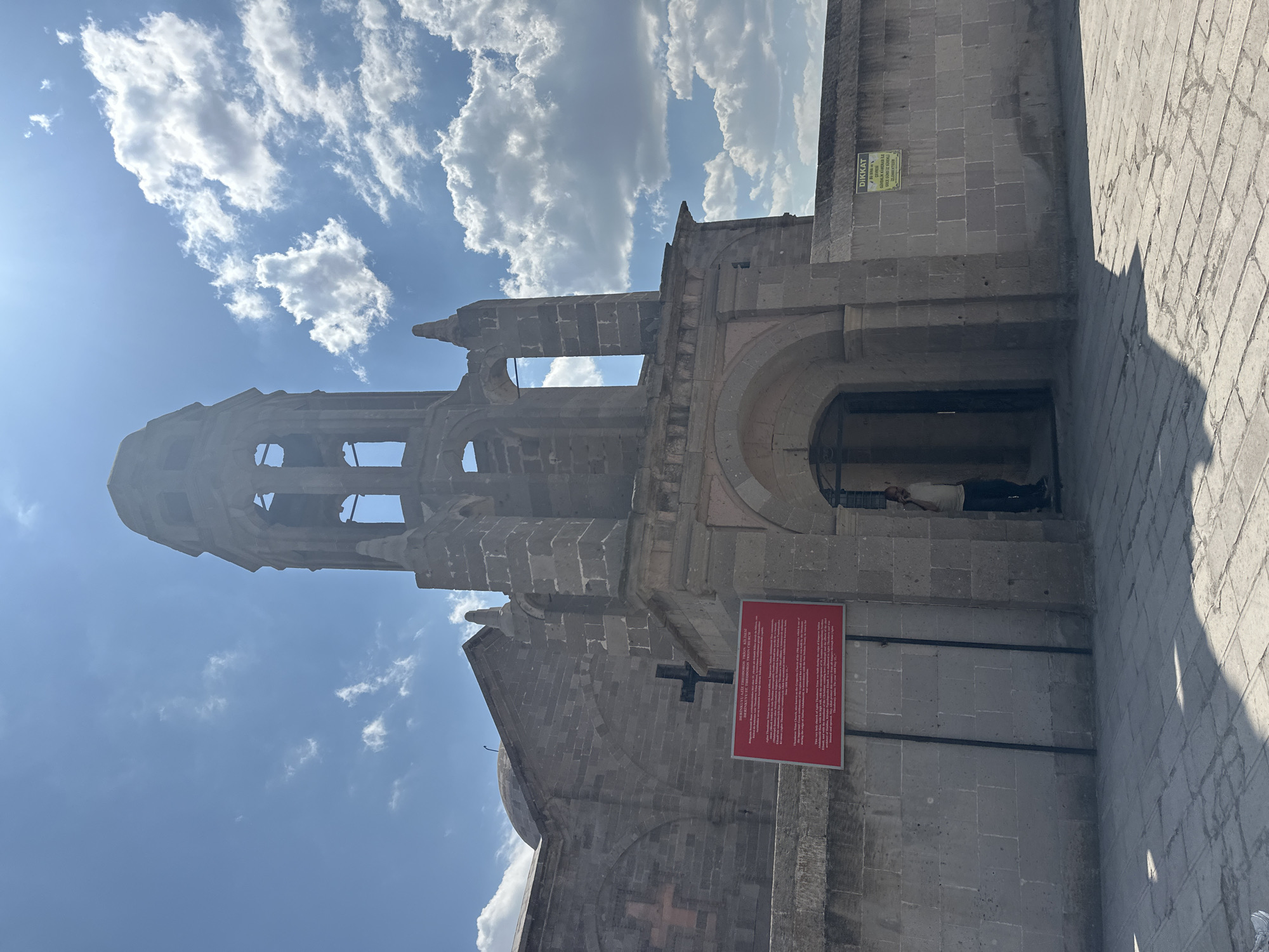 The image shows the exterior of a stone church, identified by a red sign as the Thodoreos Church in Derinkuyu, Turkey. A man is visible standing at the entrance of the church. The church has a prominent bell tower with open arches and a dome. A wall surrounds the church, and a yellow sign with the word "DİKKAT" (Danger) is affixed to it. The sky is blue with scattered white clouds. The ground is paved with stone.