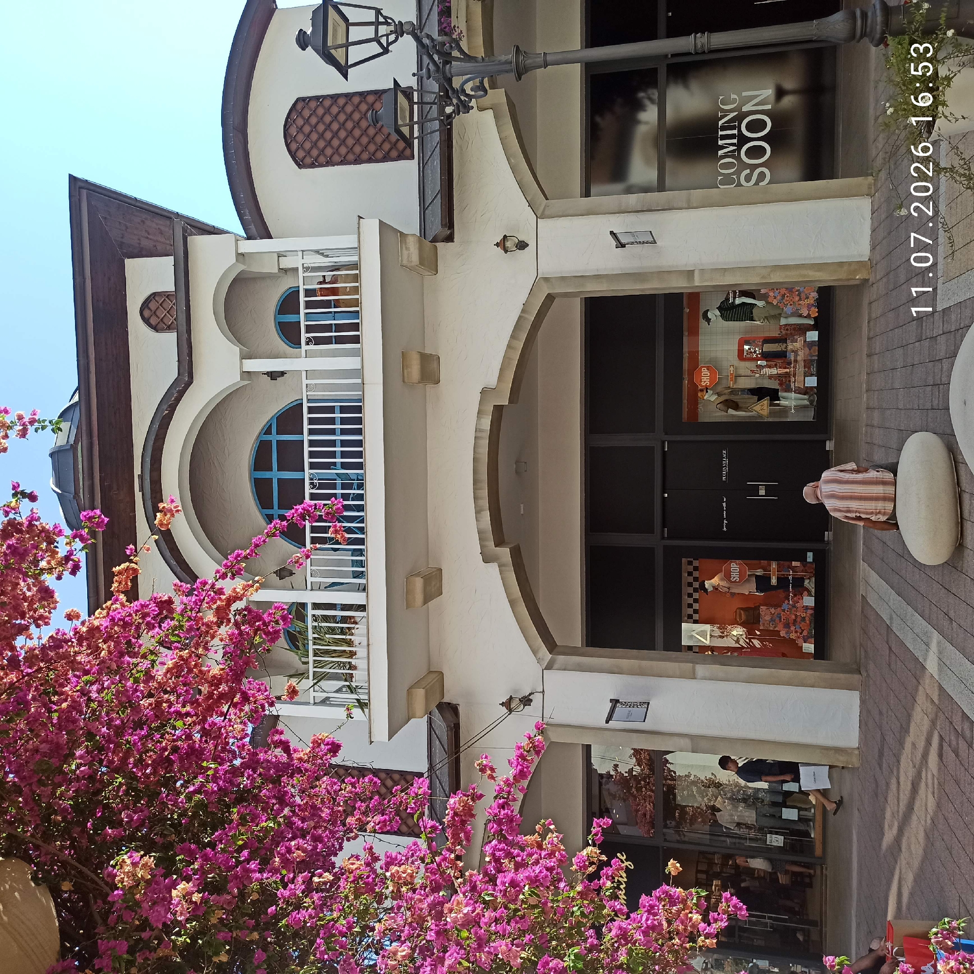 In Molfetta, Italy, a man is sitting on large stones in front of a building with storefronts. The building is white with arched entrances and a balcony on the second floor. Pink flowers are in bloom on a tree to the left of the building, and a lamppost stands to the right. The storefront windows display mannequins and signage for shops. A "COMING SOON" sign is visible on one of the windows. The ground is paved with stones. The timestamp "11.07.2026 16:53" is overlaid on the bottom right of the image.