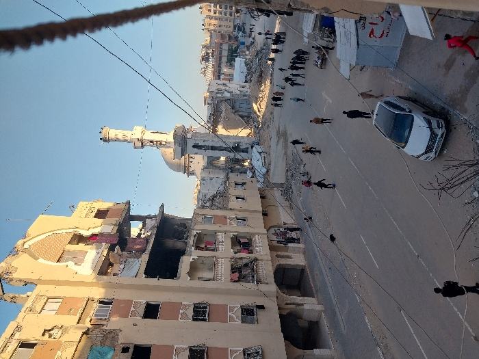 The image captures an aerial view of a street scene showing significant urban damage, likely in a residential area. The perspective is taken from a slightly elevated position, looking down onto the street and surrounding buildings.
In the background, a prominent mosque stands out with its distinct minaret and dome, appearing relatively intact compared to the adjacent structures. To the left of the mosque, a large multi-story building is severely damaged, with large sections of its facade missing, exposing internal rooms. Some areas of this building appear charred or burned, and various items, including what looks like laundry, are visible hanging from the exposed interior. Debris and rubble are scattered around the base of this damaged building.
The paved street runs through the middle ground, with a white sedan (possibly a Hyundai) parked or stopped on the right side, facing away from the viewer. Further down the street on the left, another white vehicle, possibly a van, is partially visible. Numerous individuals are seen on the street and sidewalks. A larger group of people is gathered on the left side of the street, near what appears to be more rubble or makeshift structures. Other people are walking individually along the street, navigating through the scene of destruction. One person in a vibrant red outfit is visible on the far right side of the image.
The ground surrounding the street and buildings is littered with debris, including twisted rebar and remnants of demolished structures. Overhead power or communication lines crisscross the sky, with a thick, coiled rope-like object also prominent in the immediate foreground, suggesting the photo was taken from a balcony or similar structure.
The sky is clear blue, indicating good weather and daytime conditions. On a temporary structure behind the white car, there is a white sign with red Arabic script, though the full text is not clearly decipherable; part of it reads "الر" (Al-), possibly indicating a business, organization, or temporary setup. The overall scene conveys a sense of aftermath, with residents present amidst the widespread damage.
