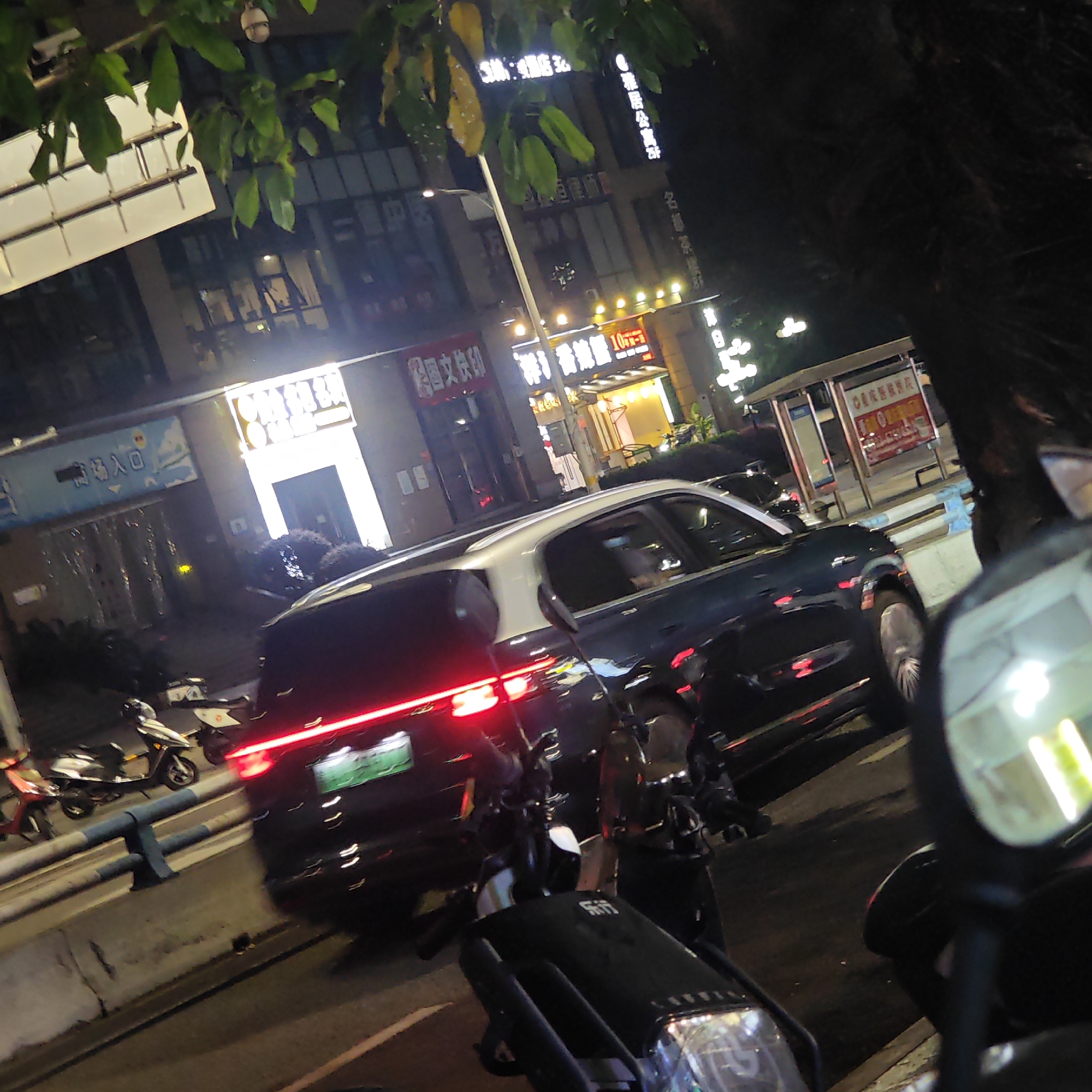 The photograph, taken at night, captures a street scene in the Yuzhong District of Chongqing, China. The image focuses on a two-tone (dark/white) car traveling on the street, its taillights illuminated. Several motorcycles are parked in the foreground, with the perspective partially obscured by the motorcycle's mirror on the right. Behind the car, buildings with lit signs in Chinese characters line the street. One sign reads "文" with other visible characters. The "雅居公寓" (Yaju Apartment) sign suggests the presence of residential buildings. The street lighting is a combination of lamps and storefront lights. There are tree branches and leaves visible in the top left corner of the frame, indicating trees along the street. A bus stop shelter is visible in the background on the right.