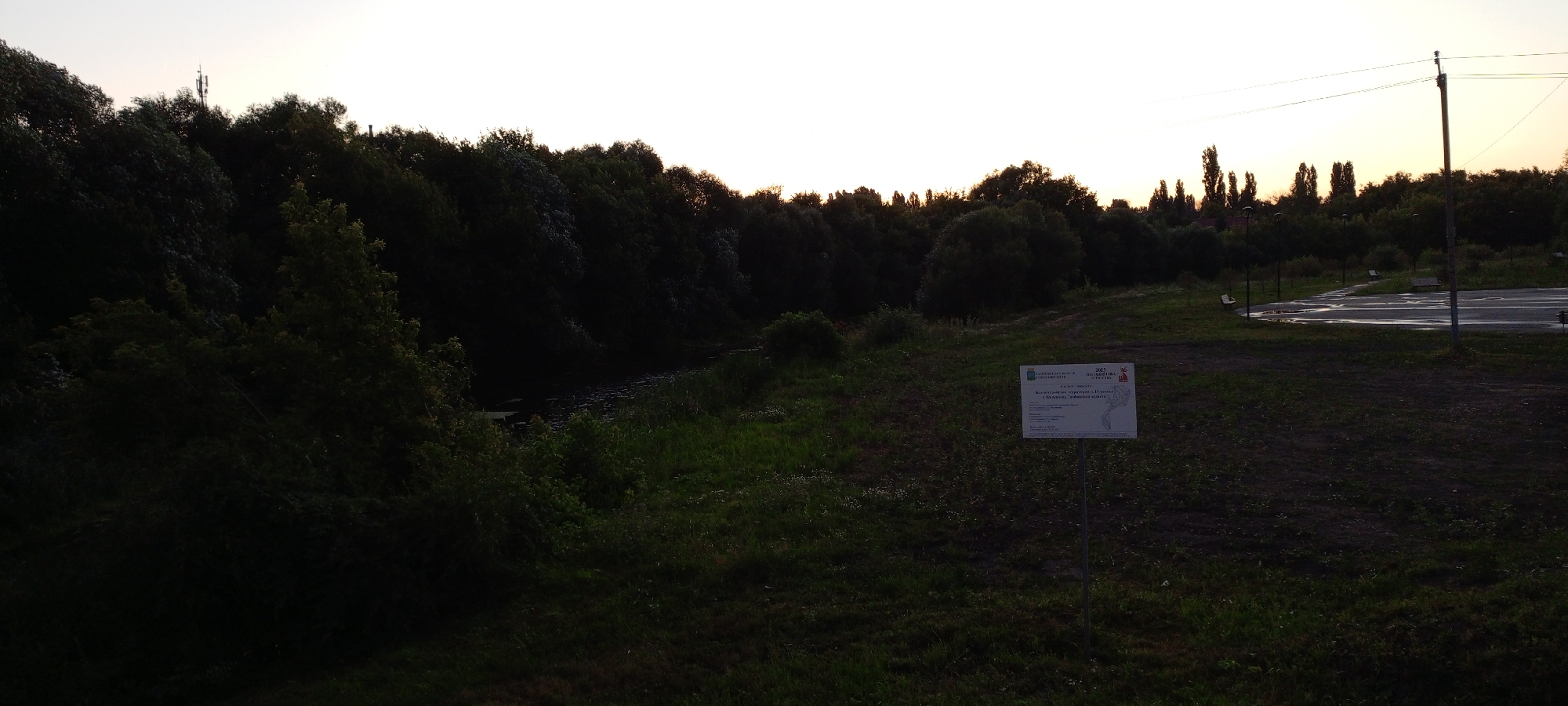 The image depicts a natural landscape at dusk, likely in Kirsanov, Russia, featuring a tree-lined riverbank with dense foliage. In the foreground, a sign is planted in the grass, displaying text and a map. Beyond the river, a line of trees stretches across the horizon, silhouetted against a pale, possibly overcast sky. A road or paved area is visible in the background, with a light pole and some benches. The overall scene is serene and natural, with signs of human presence in the form of the sign and paved area.