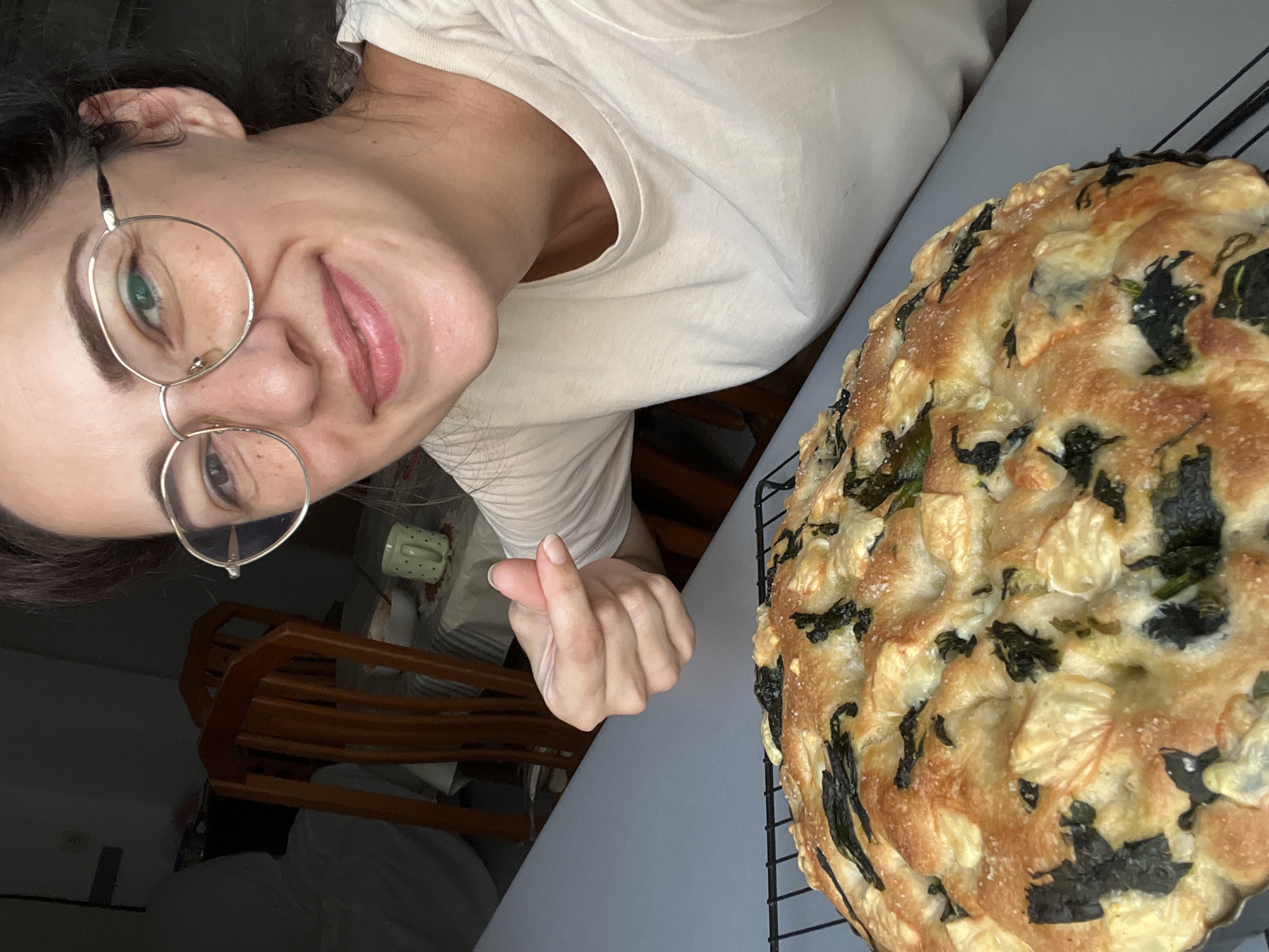 A woman with dark hair and glasses is taking a selfie in a home environment. She is smiling and making a heart shape with her fingers. In the foreground, there is a golden-brown focaccia bread with green toppings on a wire rack. The background shows wooden chairs and a table covered with a striped tablecloth. The scene is well-lit, highlighting the woman and the bread.