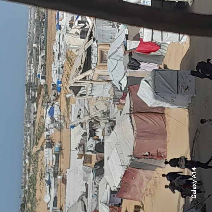 The image displays a densely populated displacement camp in Khan Yunis, Palestinian Territory, under a clear, light blue sky. The terrain consists of sandy, unpaved ground. Numerous makeshift shelters dominate the scene, constructed from a variety of materials including white tarpaulins, red and dark fabrics, and what appear to be salvaged building components like wood and corrugated metal sheets. Many shelters show signs of damage or wear, with some having collapsed or torn roofs. In the mid-ground, a black water tank is visible on top of one structure, and clothes are hanging to dry outside several shelters, including red and dark-colored garments. A stack of white rectangular objects is present near the bottom right. In the foreground, a group of approximately four to five individuals, mostly adult males, walk across the sandy ground from left to right, with one person appearing to carry an item on their head. A lone child is visible walking further into the scene. Distant structures extend to the horizon, which features some sparse vegetation or low hills. The bottom-left corner of the image contains a "Galaxy A14" watermark.