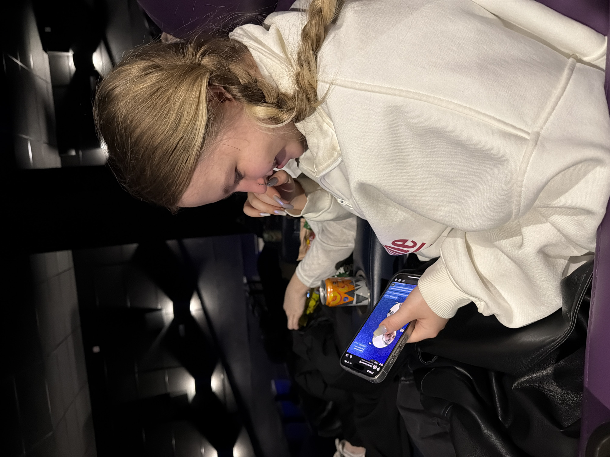 A young woman with blonde hair styled in braids is seated in a dark environment, likely a movie theater, based on the seating visible on the left side. She is wearing a white hoodie with red lettering on the sleeve, and she is holding a smartphone in her right hand. The phone's screen is illuminated, showing what appears to be a social media profile with circular headshots. With her left hand, she appears to be touching her nose or mouth. An open can of juice or soda is visible next to her left arm. There is black leather clothing or a bag in her lap. The location is described as Penza, Russia.