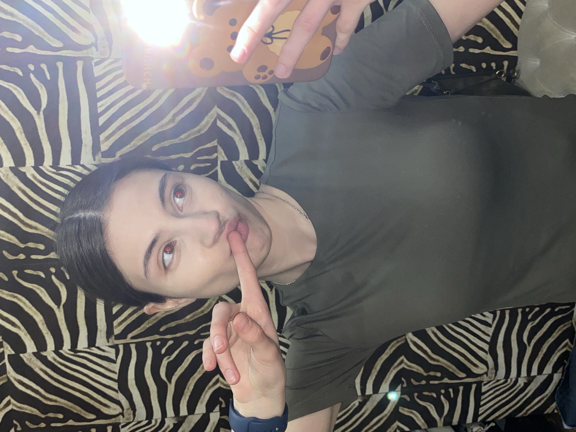 A young person is shown in a selfie, lying on a surface covered with a distinctive black and off-white zebra-print fabric, possibly a bed or couch. The individual holds a smartphone with a brown, cartoon bear-shaped case above their face, which emits a bright light, creating a strong lens flare in the upper left of the image.
The subject wears a plain olive green short-sleeved top and a silver-colored necklace. A blue smartwatch or fitness tracker is visible on their left wrist. Their dark hair is pulled back, and they look directly at the camera with wide, reddish-orange eyes. A finger from their right hand is playfully pressed against their lips in a "shushing" gesture, conveying a direct and possibly secretive or playful emotion.
Faint, mirrored Russian text, "Я МЫСЛЮ" (meaning "I THINK"), is partially visible on the phone case, obscured slightly by the lens flare. The setting appears to be indoors, likely in a dimly lit room, as indicated by the primary light source originating from the phone. The overall atmosphere is casual and intimate. The location context is Votkinsk, Russia.