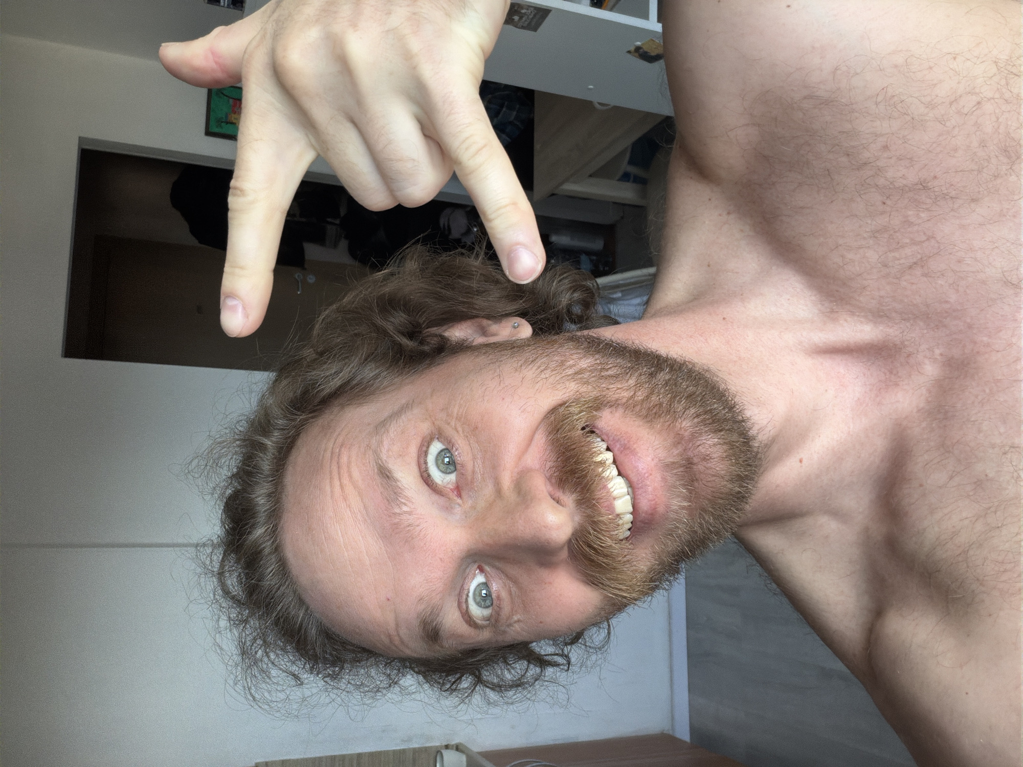 A bearded, fair-skinned man with wavy brown hair is photographed from a low angle, making a hand gesture above his head. The man's chest is bare, and he is smiling, revealing his teeth. He has light-colored eyes and appears to be indoors. The background includes a partially visible doorway and shelving. There is no information that would indicate the location of the photo is in the city of Saint Petersburg, Russia.