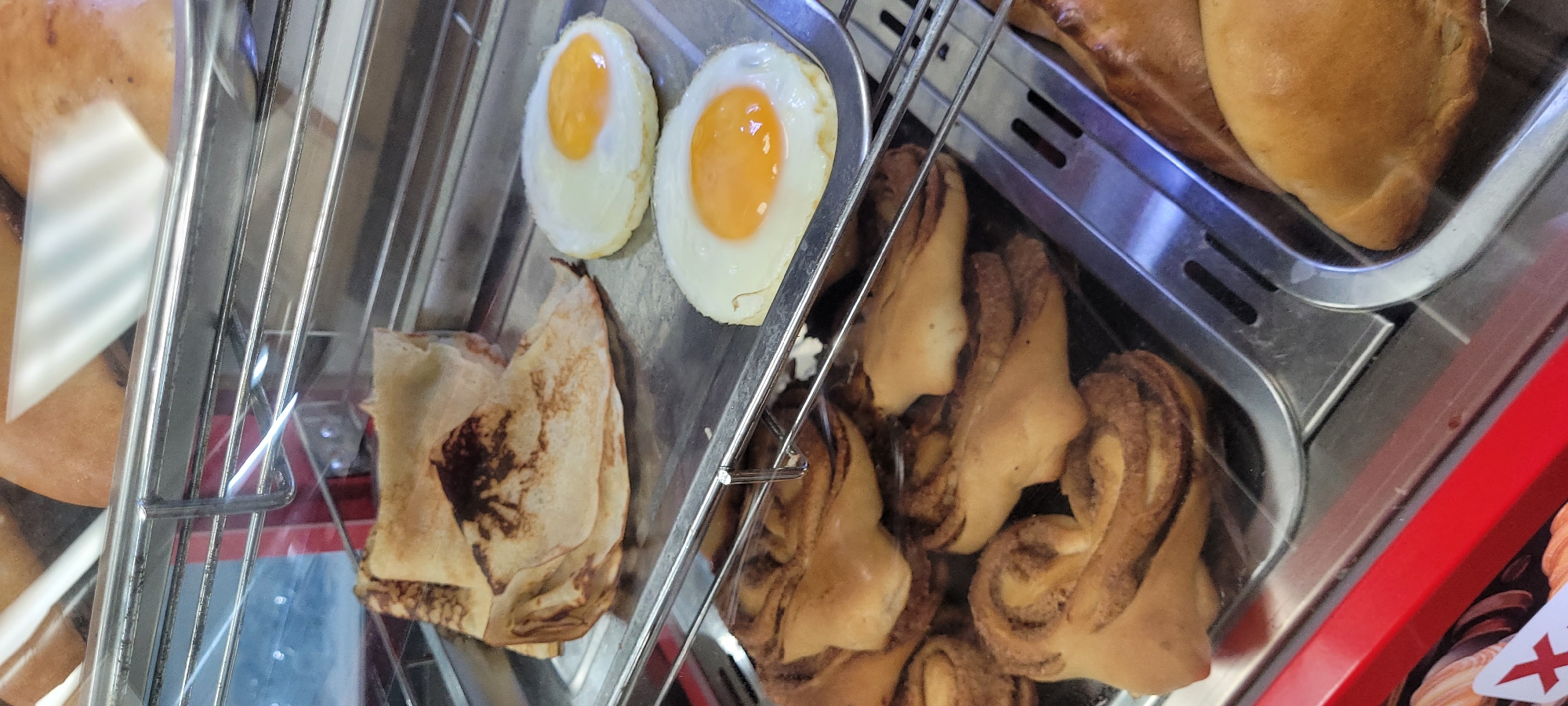 The image depicts a display case filled with various food items. On the top left tray, there are two fried eggs alongside a folded pancake. The next tray contains a selection of pastries, likely cinnamon rolls or a similar baked good. The tray on the right holds what appears to be empanadas or similar savory pastries. The scene is likely from a deli, bakery, or café in an unspecified location in the city of Unknown, Unknown. The focus is on the food items displayed for sale. There are no people visible in the image.
