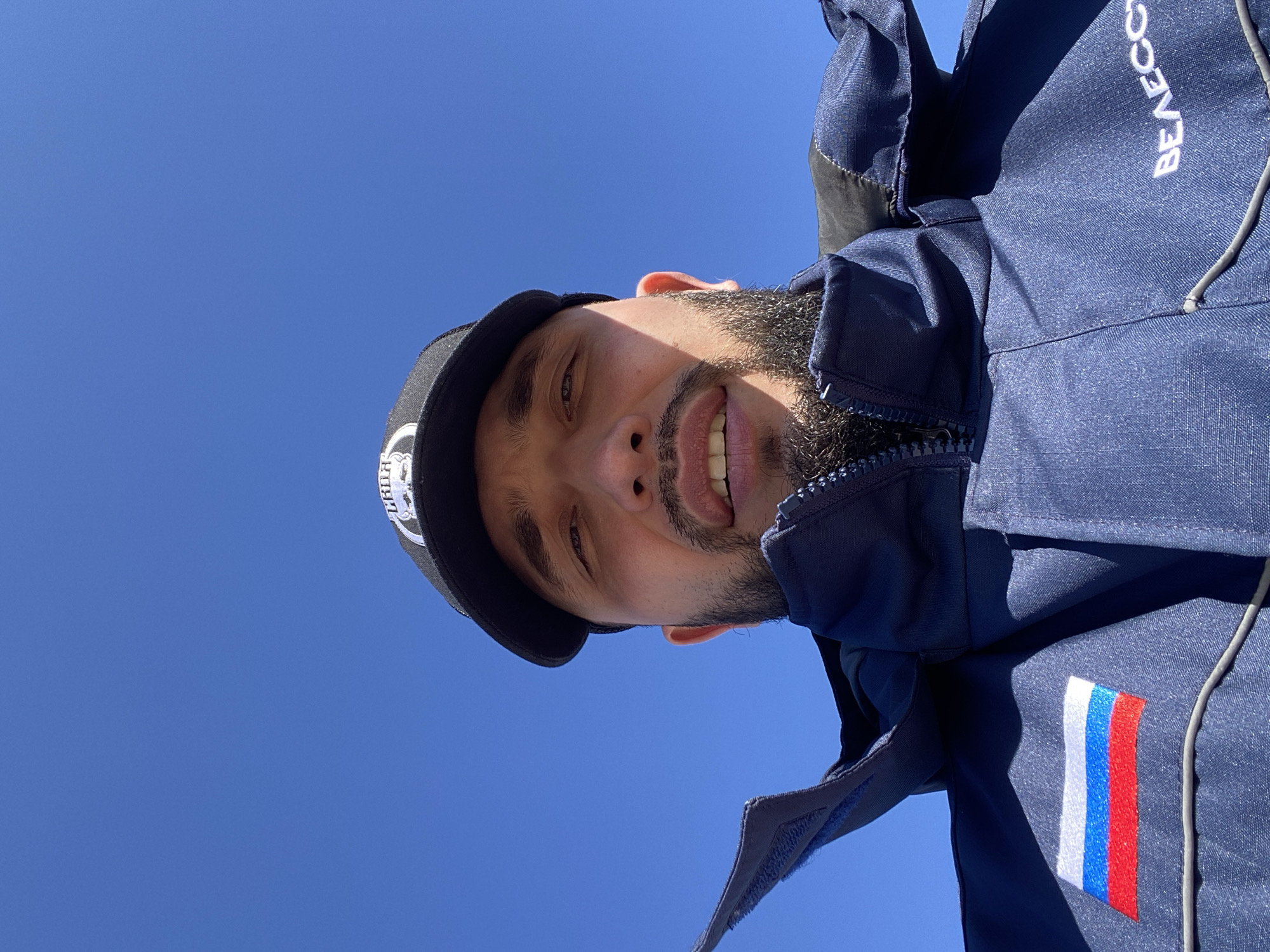 A low-angle, upside-down portrait features a man with a beard and a warm smile against a clear, bright blue sky, indicating sunny weather during the day. He is wearing a dark blue jacket and a black baseball cap. The jacket has a zippered front and a high collar. White text, "BEAECC," is visible vertically on the jacket's upper right side. On the man's left arm, a rectangular patch with the Russian flag (white, blue, and red horizontal stripes) is prominently displayed. The cap has a logo on the front, partially obscured, with "BLR" or similar letters visible. The man's face is partly shadowed, suggesting strong overhead sunlight. His expression is friendly and open. The overall setting is an outdoor location in Luzhitsy, Russia.