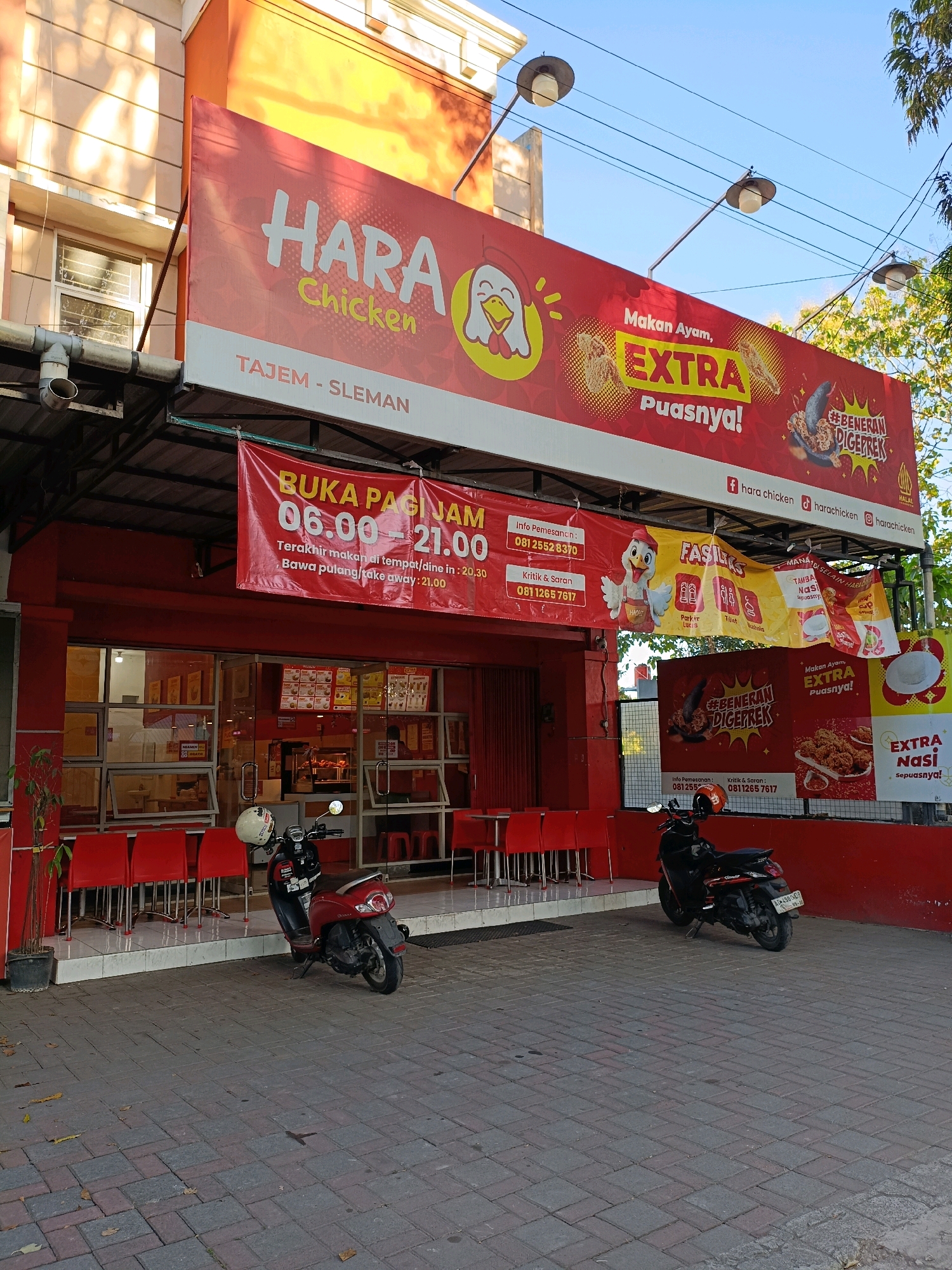 This is a medium shot, eye-level, outdoor photograph of a HARA Chicken restaurant in Ngemplak, Indonesia. The building facade is painted in bright orange and white, with a large red banner above the entrance. The banner features the restaurant's name, "HARA Chicken," in white stylized lettering, and text in Indonesian advertising fried chicken. Below this, another red banner with white text indicates the restaurant's operating hours are from 06:00 to 21:00, with the last order for dine-in at 20:30 and for takeaway at 21:00.
The restaurant entrance is glass-fronted, revealing a brightly lit interior with red chairs and tables. To the left of the entrance, a potted plant stands near a row of red chairs. To the right of the entrance, two scooters are parked on a paved walkway. The scooter in the foreground is red and white, with a white helmet resting on its handlebars. The scooter behind it is black. The walkway is made of interlocking brick pavers. In the background, trees and telephone wires are visible against a clear blue sky. The lighting suggests it is daytime.