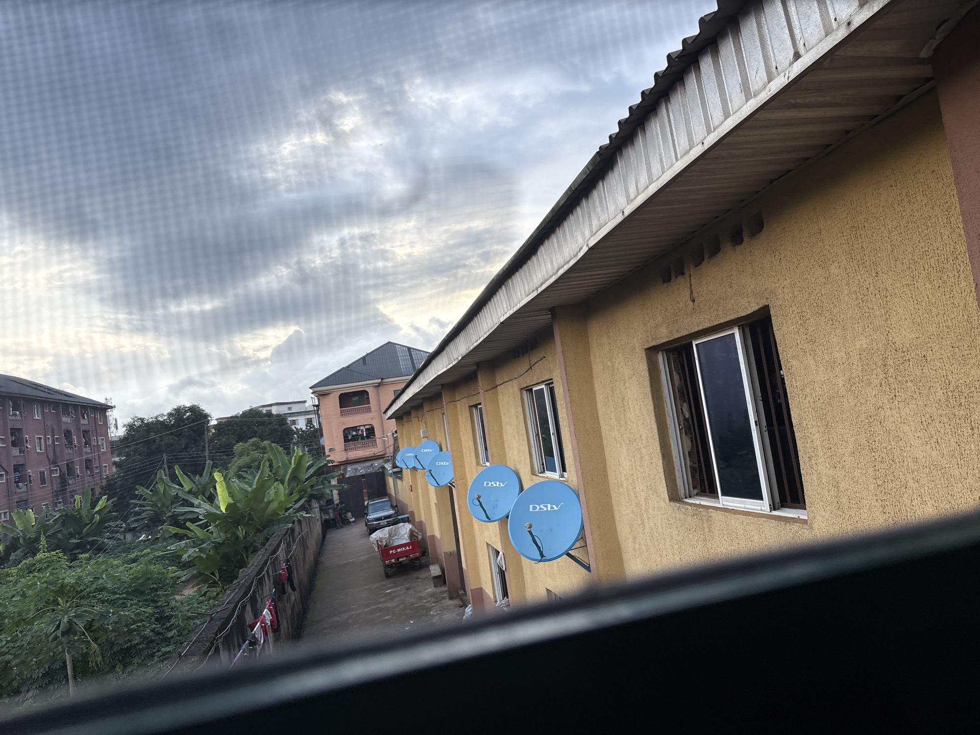 The image depicts a narrow alleyway in Onitsha, Nigeria, bordered by buildings and lush vegetation. On the right, a yellow building with a corrugated metal roof overhang has multiple blue satellite dishes mounted on its exterior wall. A window is visible on this building. Across the alley, a concrete wall separates the buildings from a dense area of banana trees and other greenery. Further in the background, other multi-story buildings can be seen, including a pink one with balconies. A small red truck with a canopy is parked in the alley, along with other vehicles. Laundry hangs on a clothesline along the wall. The sky above is overcast with dramatic cloud formations. The overall scene suggests a residential or mixed-use area in a tropical urban environment.