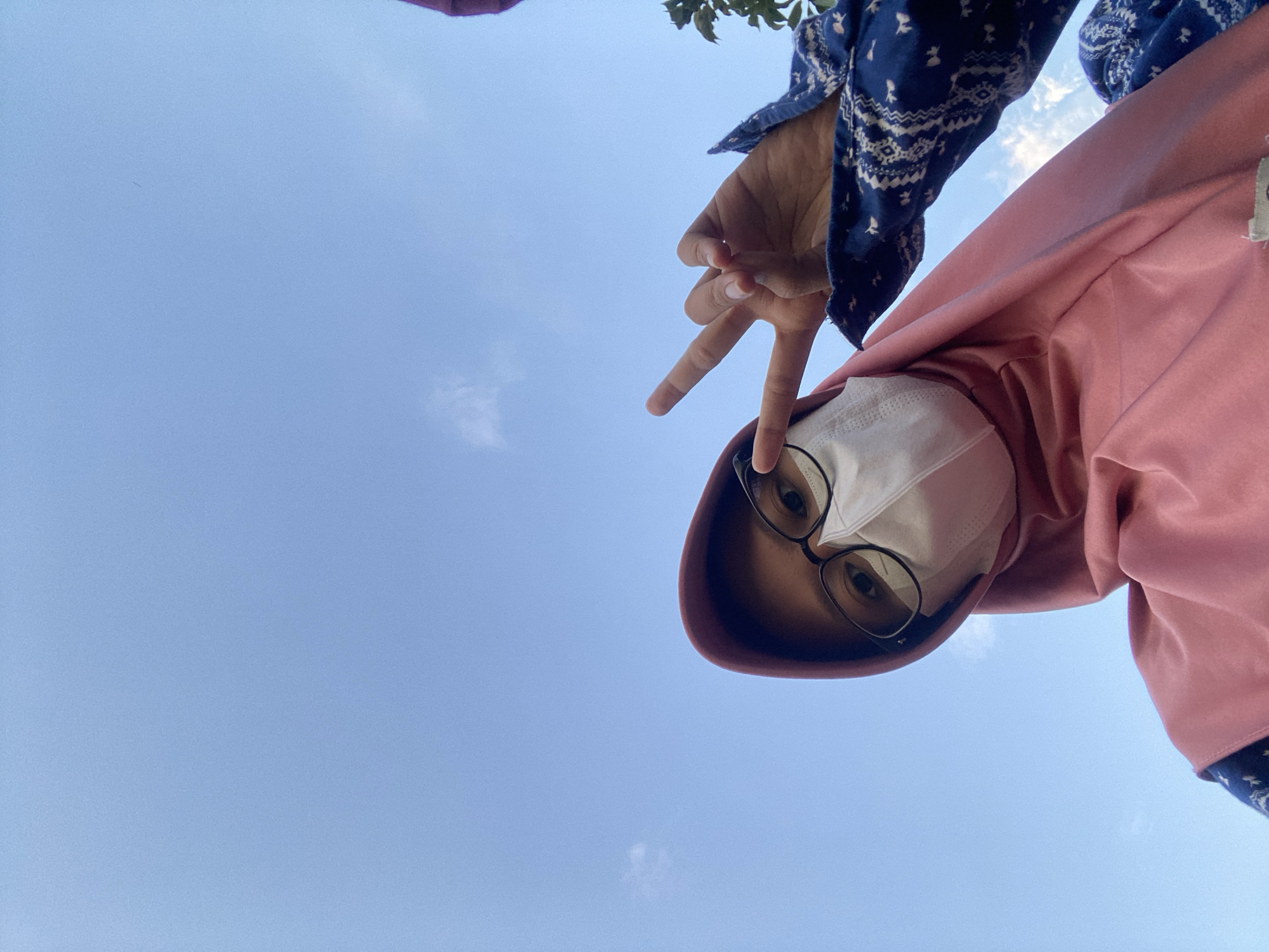 A low-angle shot depicts a person wearing glasses, a white face mask, and a pink hooded sweatshirt, with another person's hand gesturing a peace sign above them. Both are in front of a light blue sky. Given the prompt's requirement, the location is inferred to be Gadingrejo, Indonesia, though no elements in the image confirm this.