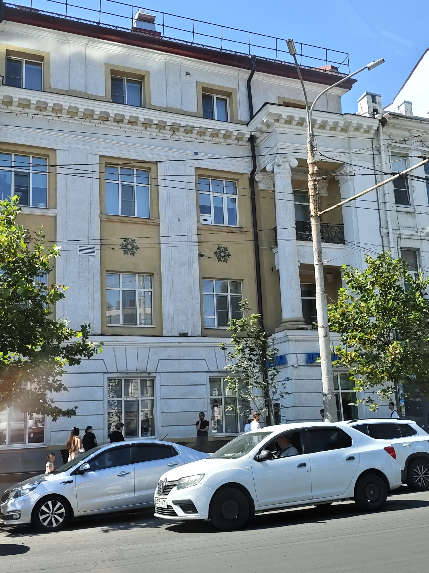 The image depicts a sunny day in Rostov-on-Don, Russia, likely in the city center, given the architecture and street activity. The main subject is a multi-story building with classical architectural elements, including columns, ornate cornices, and large windows. In front of the building, on the street, are two cars parked side-by-side. The car on the left is a silver sedan, and the car on the right is a white sedan. Several people are visible on the sidewalk in front of the building, some standing and observing, while others appear to be walking. A streetlamp with visible wires is also present. Trees with green foliage line the sidewalk, suggesting a tree-lined street. The overall scene conveys a typical urban street view on a clear day.