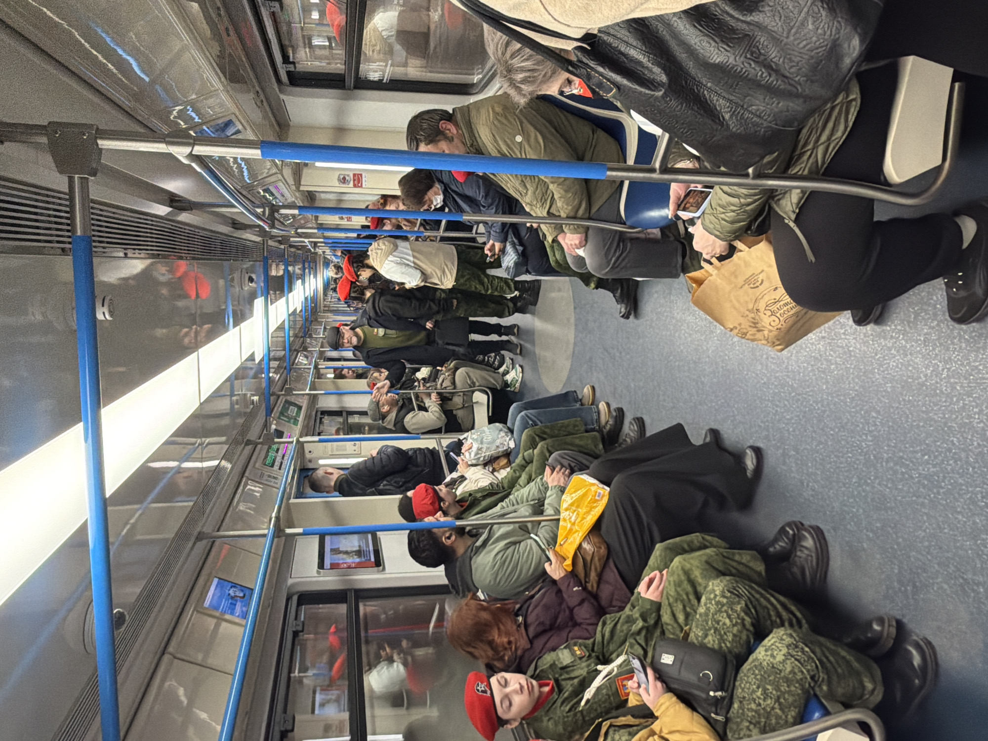 The image captures the interior of a subway train car, oriented sideways. Many passengers, including men and women of varying ages, are seated along the length of the carriage, with some standing or leaning against poles. The scene suggests a typical commuting period, with people generally calm and engaged in personal activities like using their phones, resting, or observing their surroundings.
**Subjects:** The train is occupied by a diverse group of individuals. Prominently in the foreground is a young person wearing a military-style camouflage uniform and a distinct red beret. Other passengers are dressed in civilian attire, including various jackets and coats suitable for cooler weather.
**Objects:** The train car features a contemporary design with blue handrails and support poles, grey and blue seating, and reflective surfaces on the walls and ceiling. Digital display screens are integrated into the interior walls, one of which shows a landscape image and some text. Passengers carry personal items like phones, handbags, and a distinctive brown paper bag.
**Activity or Event:** The primary activity is public transportation. Passengers are traveling, either seated or standing. Many are looking at their mobile phones, while others appear to be resting or observing the train's interior.
**Notable Details:** The lighting is primarily artificial from within the train car. Clothing worn by passengers suggests cooler temperatures. The presence of a person in a military-style uniform with a red beret stands out among the general commuters. The overall ambiance is routine and unhurried.
**Visible Text:** On the brown paper bag held by a passenger, the Cyrillic text "Золотое России" (Zolotoye Rossii), which translates to "Golden Russia," is clearly visible. One digital screen displays "4K" along with some smaller, unreadable Cyrillic text. Other small signs and displays, likely in Cyrillic, are present but too indistinct to decipher.