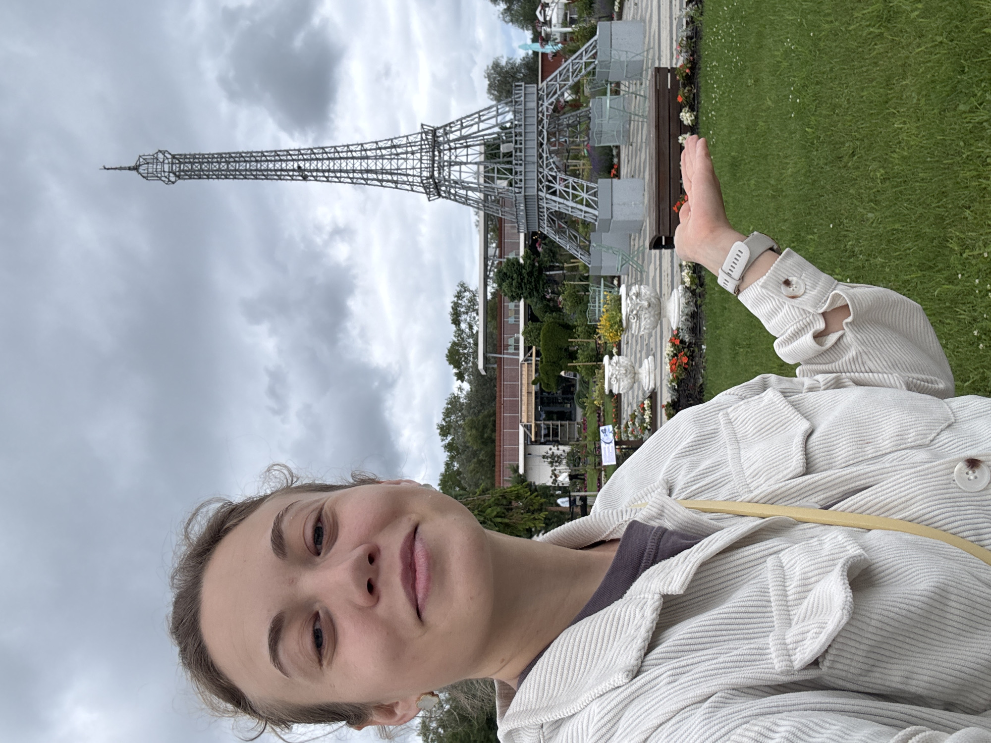 A young woman stands in a park, holding her hand out towards the camera with her palm up. She is wearing a white corduroy jacket and a dark t-shirt. In the background, a miniature replica of the Eiffel Tower stands tall against a cloudy sky. The park features manicured lawns, flowerbeds, and outdoor seating areas. The location is identified as Bruzdeilynas, Lithuania.