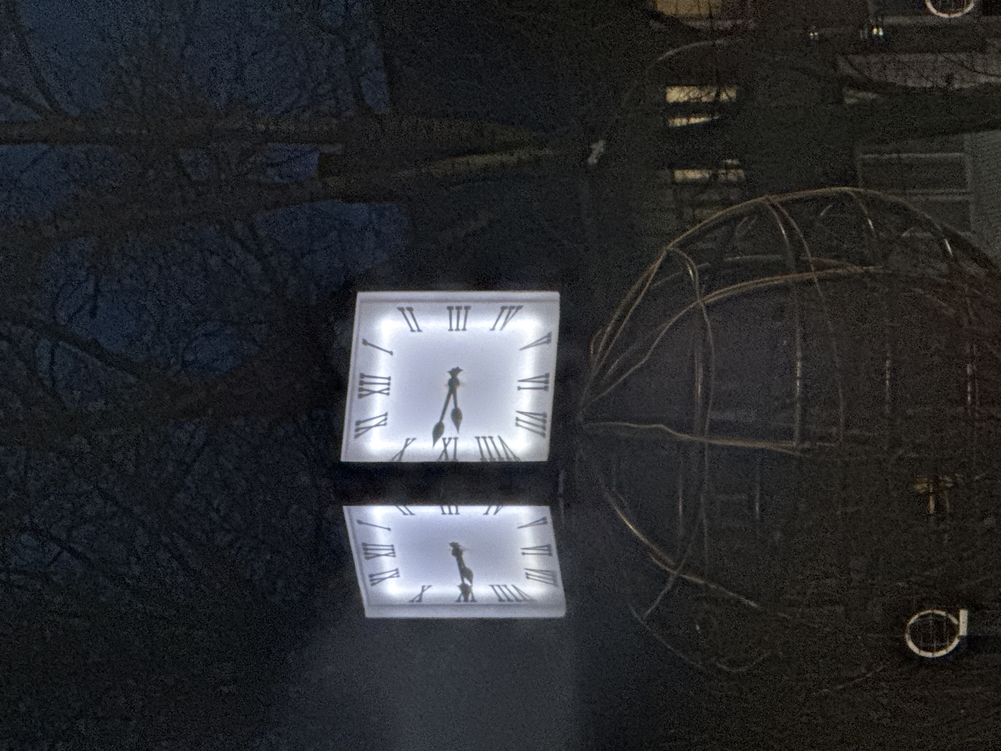 A nighttime photo depicts a square clock face illuminated with white light, reflecting off a dark surface below. The clock shows the time as approximately 8:50, indicated by the position of its hands. The reflection below mirrors the clock's face and hands. To the right of the clock and its reflection is a wire-frame globe-like structure. The environment surrounding the clock and globe is a dark, blurry mix of trees and potentially a building, suggesting an urban or park-like setting. It's in Chaikovsky, Russia.