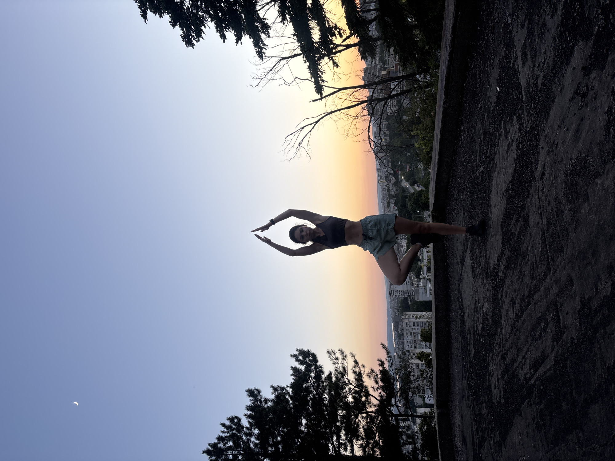 A woman in athletic wear is performing a tree pose yoga stance on a dark, rough surface. She is silhouetted against a gradient sky transitioning from a pale blue at the top to a warm orange and pink near the horizon, indicative of either sunrise or sunset. A crescent moon is visible in the upper left quadrant of the sky. The background displays a cityscape with buildings and trees, suggesting an urban environment. The woman's arms are extended upwards, and her right foot is placed on the inner thigh of her left leg, with her left leg supporting her weight. In the foreground, the textured, dark ground appears to be a rooftop or an elevated platform. The overall scene captures a moment of calm and physical activity against a picturesque sky with an urban backdrop. The location is Varna, Bulgaria, given the specific architectural style and urban planning visible in the cityscape and the presence of trees common in the region.