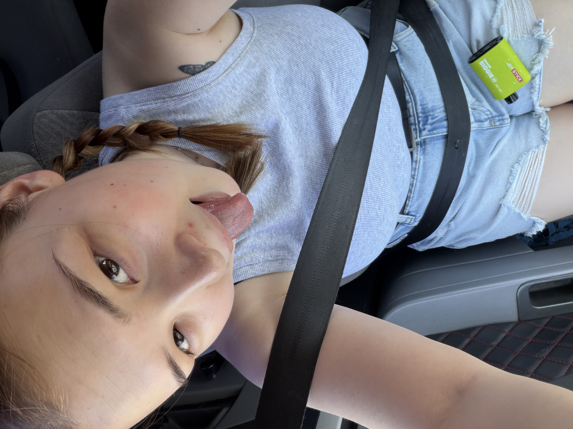 A young woman is in the driver's seat of a vehicle. She is wearing a light blue shirt and ripped denim shorts, with a seatbelt fastened across her body. Her hair is styled in pigtail braids. She is sticking her tongue out and looking at the camera. A "Grand Biggie Joy Stick" vape pen is visible in her lap. The interior of the vehicle includes a grey center console with red stitching. The location is presumably in the city of Chaikovsky, Russia, although there are no specific indicators within the frame.
