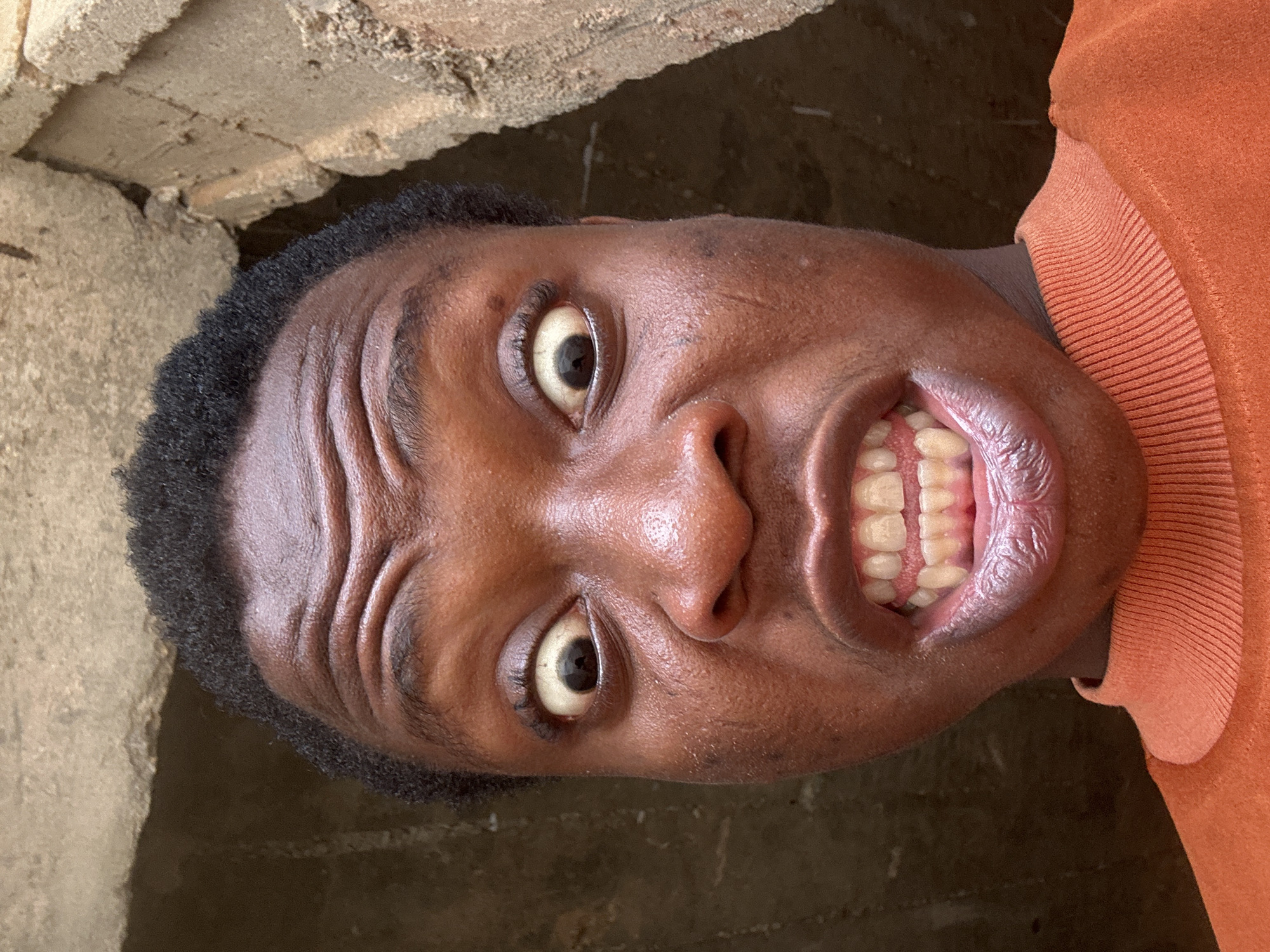 The image shows a close-up, upside-down view of a Black male's face. The individual is positioned near a concrete wall or structure. His mouth is open, revealing his teeth. He is wearing an orange sweater. The environment appears to be a dimly lit, possibly urban setting. The location is unknown.