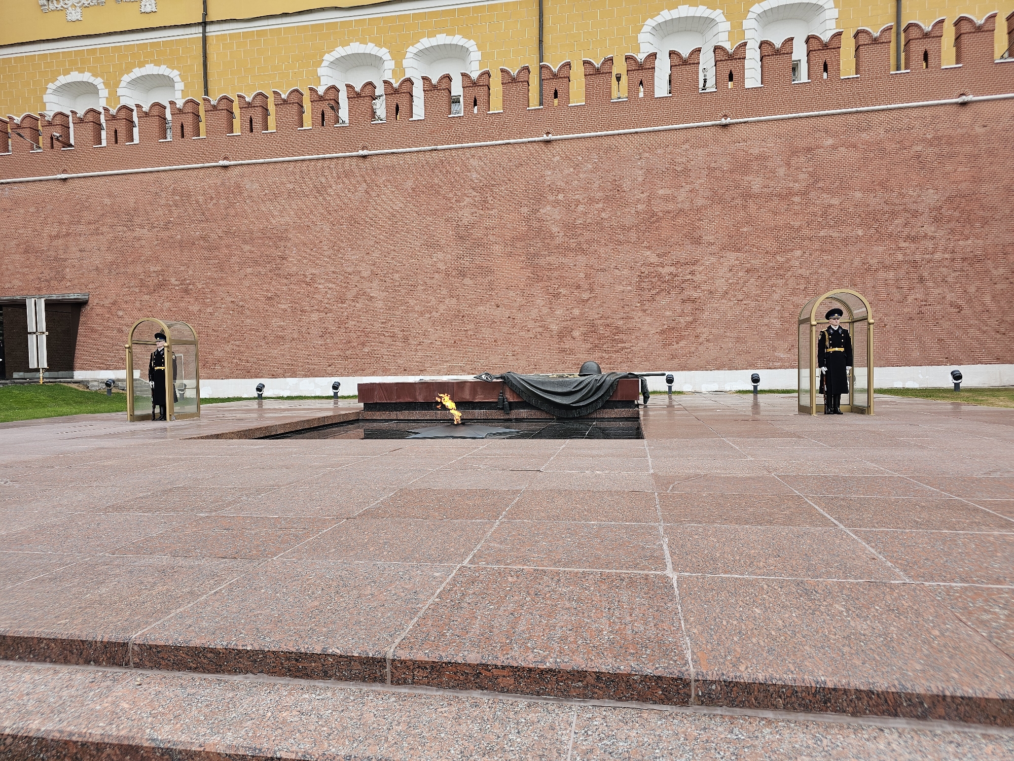 The image depicts the Tomb of the Unknown Soldier in Moscow, Russia. A red brick wall, part of the Kremlin, forms the backdrop. At the center of the scene is a dark granite monument with an eternal flame burning in front. A dark cloth partially covers the monument. Flanking the monument on either side are two honor guards standing motionless in golden-framed glass enclosures. The foreground is a large granite plaza with visible joints between the paving stones, leading to the lower edge of the frame. The day appears overcast, as suggested by the flat lighting and reflections on the plaza.
