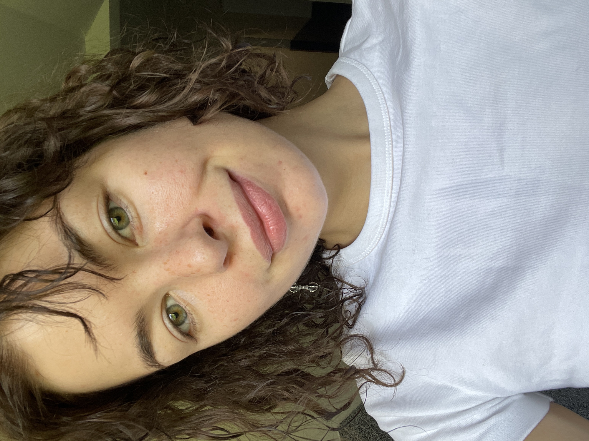 A close-up, tilted portrait features a young woman with shoulder-length, dark brown, curly hair that frames her face. She has fair skin with visible freckles across her cheeks and nose. Her eyes are a striking green and look directly at the viewer. Her eyebrows are dark and well-defined. Her lips are a light pink, slightly parted, revealing no teeth.
She is wearing a simple white ribbed t-shirt. A small, intricately designed metallic earring, resembling a vajra symbol, is visible on her right ear (viewer's left), peeking through her hair. The background is indistinct, appearing as a plain, lighter-colored surface, possibly an indoor wall or curtain, creating a soft backdrop that keeps the focus on her face.
The lighting appears natural and even, suggesting a daytime setting, though no specific light source or outdoor elements are visible. The subject has a neutral to slightly relaxed expression, with a hint of a smile. There is no visible text in the image. The location context provided is Boreti, Montenegro, but no specific geographical cues are discernible within the image itself.