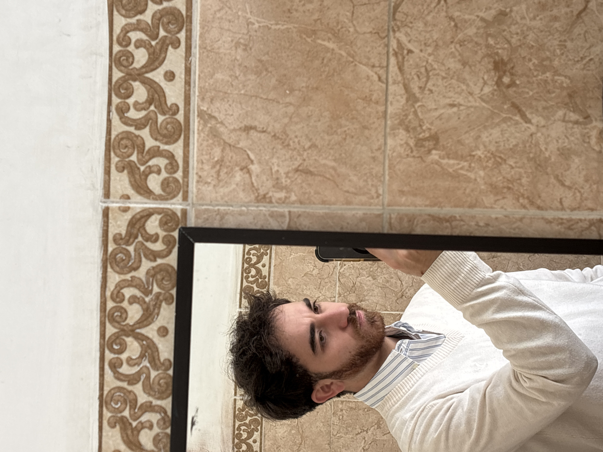 A man is taking a selfie in a mirror in what appears to be a bathroom. The wall behind the mirror has beige tiles with a decorative brown pattern. The man has dark curly hair and a reddish-brown beard. He is wearing a white sweater over a striped shirt. The location appears to be in Puebla, Mexico, based on the style of the tiles and the wall patterns, which are common in the area. The focus is on the man and his reflection in the mirror, suggesting he is the subject of the photo. He holds his phone above the frame. The environment is a typical bathroom setting.