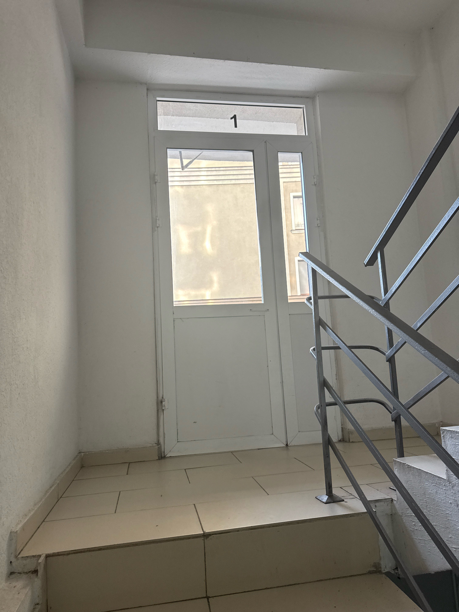 The image depicts the interior of a building's stairwell, specifically at what appears to be the first landing. A white door with a rectangular glass pane at the top is centrally located. The number "1" is visible in black above the door. To the right of the door, a metal staircase railing with a gray finish is prominent, leading upwards. The walls are painted white and appear to be textured. The floor is tiled with large, light-colored square tiles. Outside the glass pane of the door, a building with visible window frames and a muted facade is discernable. The scene suggests a typical urban apartment building environment, possibly in Chișinău, Moldova, given the context provided. There are no individuals present in the frame.