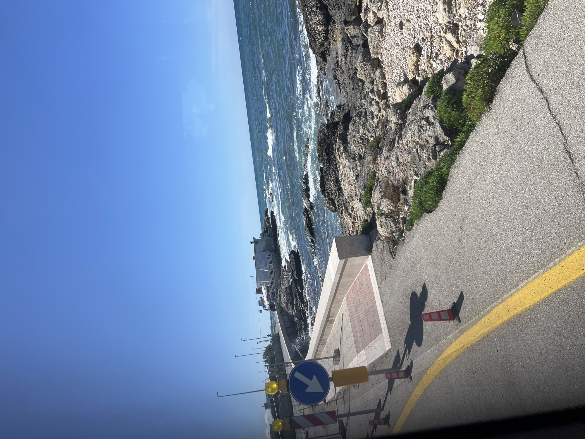 The image captures a sunny, clear day along a rocky coastline, with a road running parallel to the sea. The perspective is slightly elevated, looking down and along the curve of the road.
The top-left portion of the image is dominated by a brilliant, clear blue sky. Below the sky, to the left, the deep blue-green sea stretches towards the horizon, with visible white-capped waves breaking near the shore.
On the right side of the frame, a grey asphalt road is visible, featuring a prominent solid yellow line delineating its edge. Several traffic management elements are present on and alongside the road. A blue circular sign with a white arrow pointing diagonally downwards and to the right is a central feature, indicating a required direction. Nearby, multiple red and white striped safety barriers or cones are positioned. Yellow light fixtures on poles are also seen. Long, sharp shadows are cast across the road from these objects, suggesting bright sunshine.
Adjacent to the road, a low concrete barrier or wall separates it from the rocky shore below. This barrier includes a section with a distinct reddish-brown and lighter grey square pattern. The coastline itself is rugged and rocky, with sparse green vegetation growing in patches amidst the rocks, extending down to the water's edge. In the mid-ground, further along the coast, several white buildings are visible, possibly structures like restaurants or houses.
No people are visible in the scene. The weather is clear and sunny, indicating daytime, likely morning or early afternoon given the angle of the shadows. The scene depicts a coastal road in Bari, Italy.