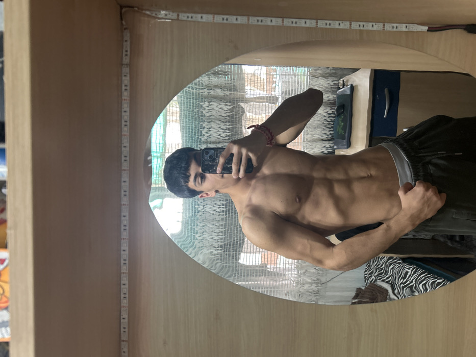 The image captures a mirror selfie taken indoors within a residential setting in Pernik, Bulgaria. A young male, approximately late teens to early twenties, is depicted shirtless, showcasing a muscular physique with defined abdominal, pectoral, and arm musculature. He holds a dark-colored smartphone in his right hand, positioned in front of his face, to capture his reflection. His left hand is placed on his hip, gently grasping the elastic waistband of his dark grey athletic pants, beneath which a light-colored undershirt waistband is visible. A red and white braided bracelet adorns his left wrist.
The subject is reflected in an arched mirror set into a light-colored wooden panel, likely part of a shelf or furniture unit. LED strip lights are affixed along the internal perimeter of this wooden frame. The mirror's reflection reveals further details of the room: a window covered by patterned sheer white curtains, offering a faint view of an exterior brick building. To the right of the reflected subject, a light-colored desk with a blue drawer is visible, upon which rests a dark mouse pad featuring a green design. To the left, a bed with black and white zebra-print bedding is partially visible. Above the wooden mirror frame, a shelf is present, containing several small items, including a multi-colored box and a blue container.