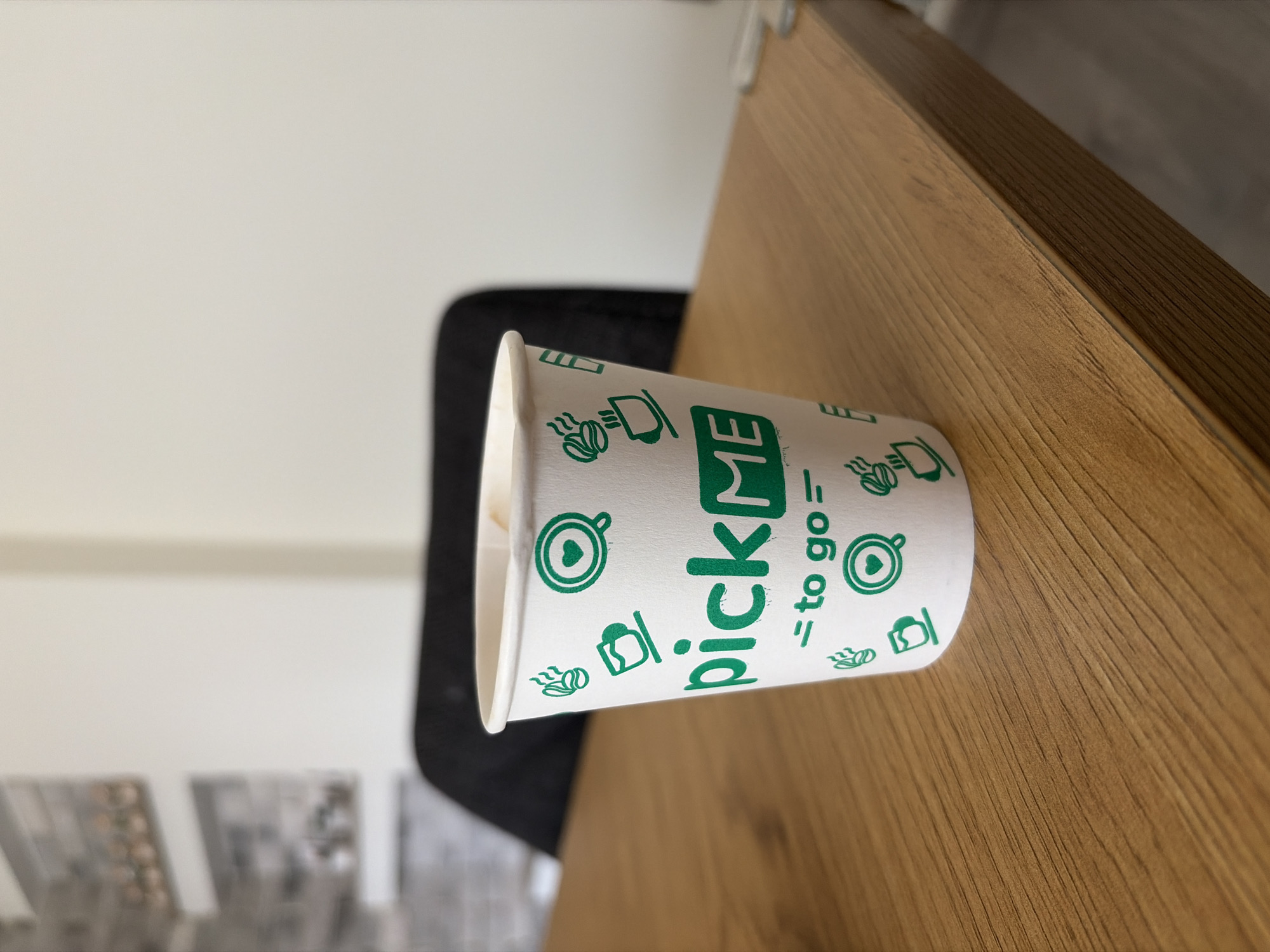 An indoor scene in Svetovrachene, Bulgaria, features a white paper cup with green branding, lying horizontally on a light brown wooden surface. The cup is the primary subject, adorned with the prominent green text "pickME" in a stylized font, followed by "= to go =". Surrounding these words are various small green icons related to beverages, including steaming coffee cups, coffee beans, and a cup with a heart design.
The wooden surface beneath the cup displays a clear grain pattern, suggesting a natural wood table or desk. In the background, a dark, indistinct object, likely a chair or other furniture, is visible against a plain, light-colored wall. Further in the blurred distance to the left, faint shapes that could be decorative tiles or framed art are discernible.
The scene is brightly and evenly lit, characteristic of daytime. There is no specific activity taking place, as the cup remains static. The image is presented sideways, reflecting its original orientation during capture. No individuals are present in the frame, and the overall mood is neutral.