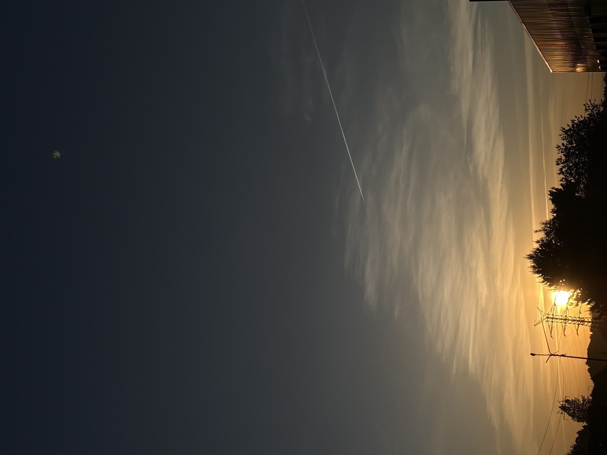 Here is a description of the image:
The image shows a sky with a dark blue area on the left and a sunrise or sunset on the right. A contrail runs diagonally across the sky. There is a small, blurry green object in the dark blue area. On the right side, trees and electrical pylons are silhouetted against the bright sky. The building is located in the city of Chaikovsky, Russia.