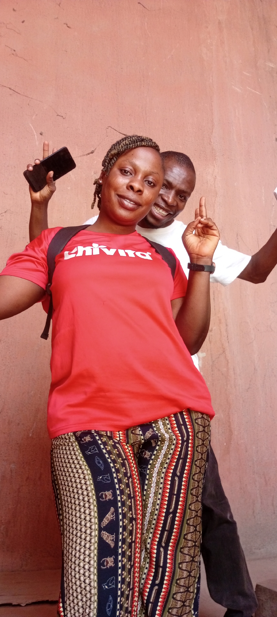 This vertical shot captures two cheerful individuals, a woman and a man, posing for a photo against a plain, textured reddish-brown wall in Fagge C, Nigeria.
The woman is in the foreground, smiling broadly at the camera. She has braided hair and is wearing a bright red t-shirt with the word "Chivita" printed in white, accompanied by a small green leaf-like logo above the 'i'. Over her shoulders, a black backpack strap is visible. Her pants feature an elaborate, multi-patterned design with vertical stripes and various motifs in colors including black, white, red, green, and gold. She holds a black smartphone in her raised left hand, making a V-sign or peace gesture with her fingers. A black watch is visible on her right wrist.
Behind and slightly to her right, the man also smiles widely, looking at the camera. He is wearing a white t-shirt and dark trousers. His right arm is raised, with his index finger pointing upwards. Both subjects exhibit happy and friendly expressions. The lighting is bright and even, suggesting it is daytime. The wall behind them shows some minor cracks and texture.