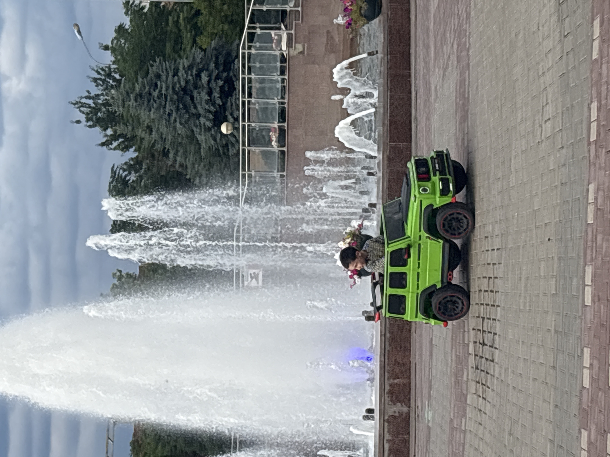 A young boy sits in a bright green toy Mercedes-Benz G-Wagon on a paved plaza. He is looking to his right, and appears to be smiling. Behind him, a large fountain sprays water high into the air, illuminated by blue lights. The plaza is paved with light gray and red brick pavers. In the background, there are trees, a street light, and a railing with planters. The scene appears to be a public park or square. This specific fountain is located in Astrakhan, Russia.