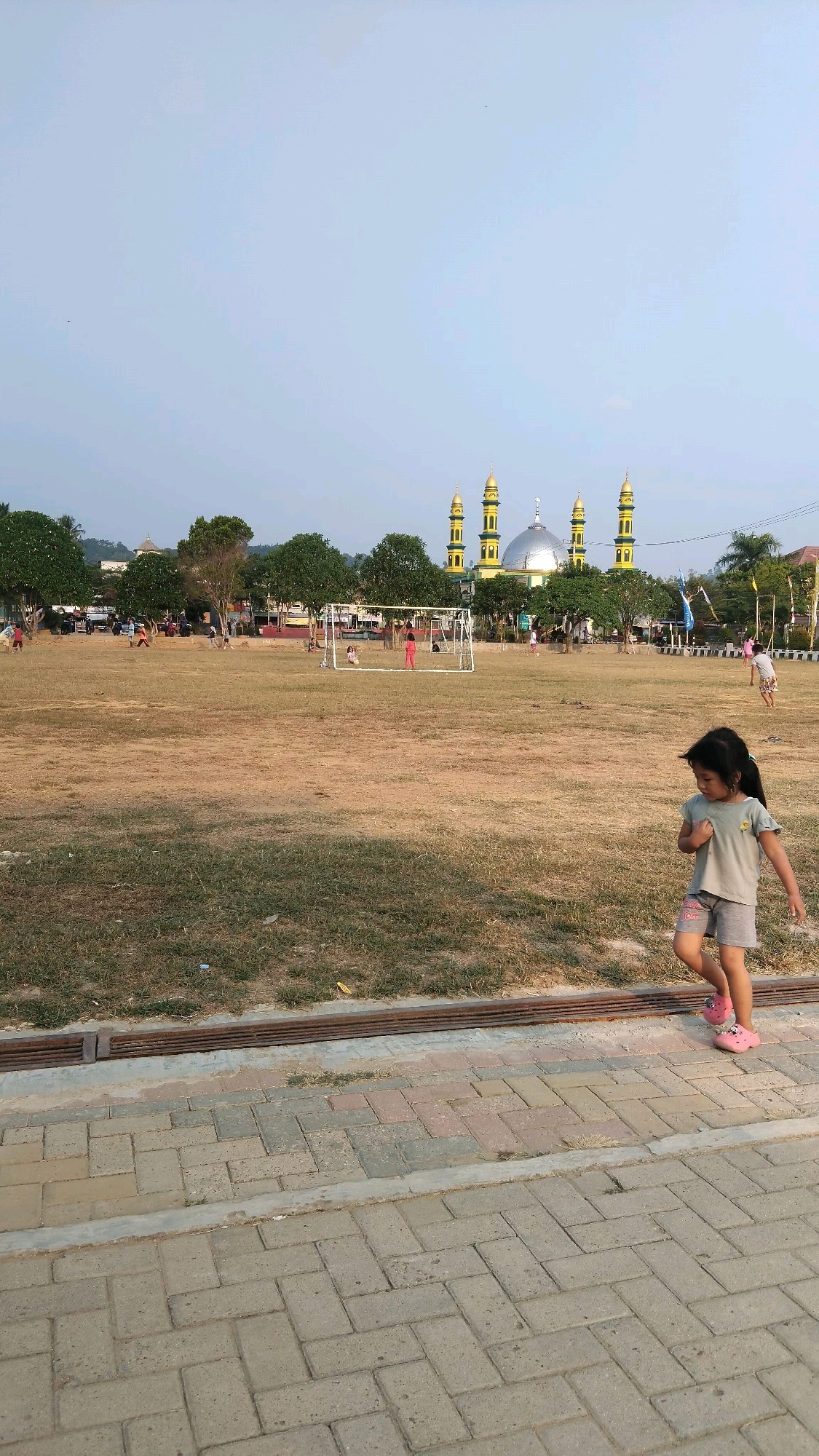 A young girl walks on a paved path in front of a dry grass field. In the background, a mosque with a dome and four minarets stands prominently. Trees and buildings are visible behind the field, and several people are scattered across the field, some playing with a soccer goal. The sky is clear and light blue. The scene appears to be set in Manunggal Jaya, Indonesia, a location characterized by its open spaces and proximity to religious structures.