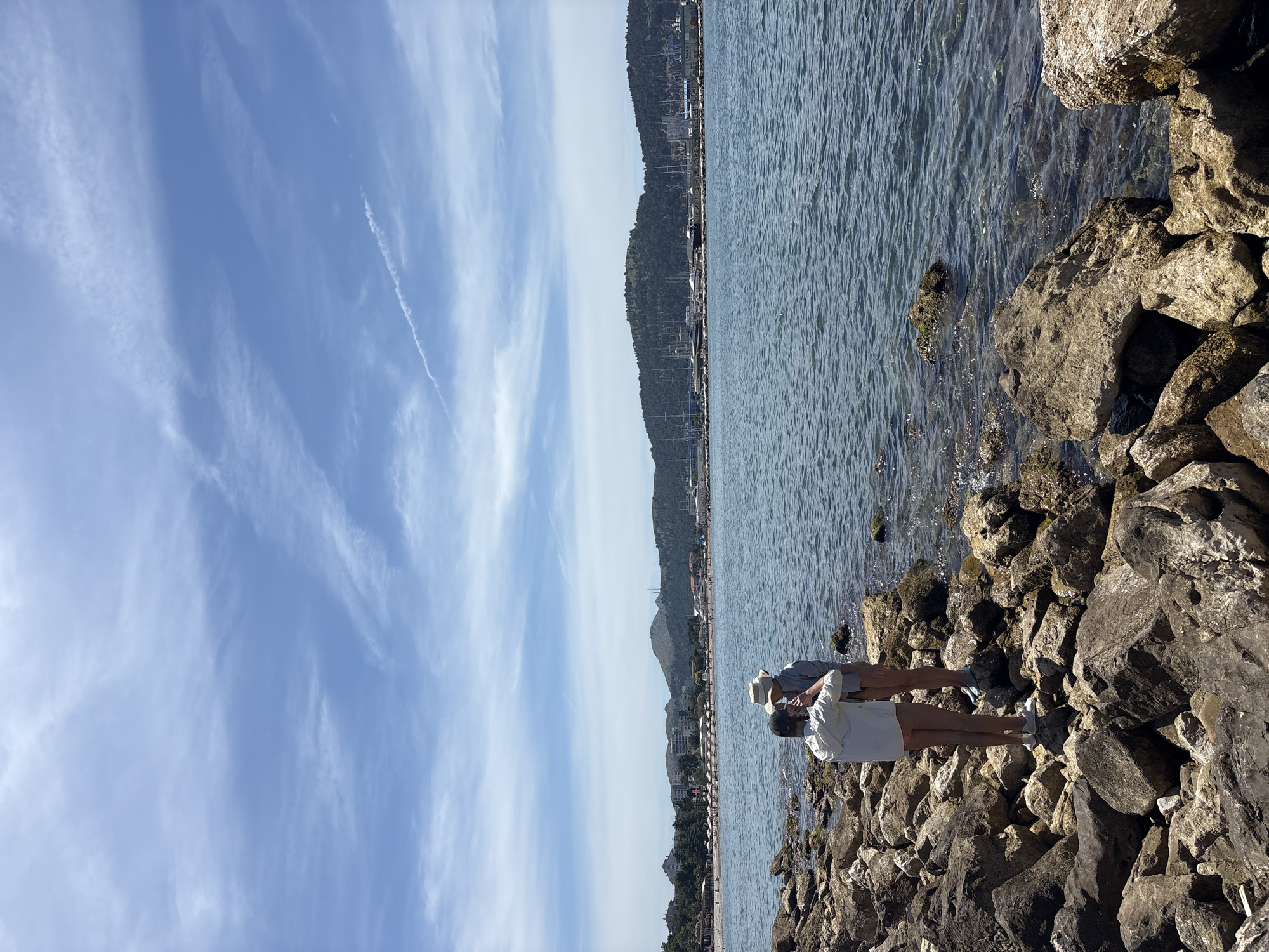 Two individuals stand on a rocky shoreline overlooking the Adriatic Sea. The person on the left, facing away from the camera, wears a light-colored hooded jacket and pink shorts. The individual on the right, also facing away, is wearing a light blue shirt and a white hat. They appear to be interacting, possibly taking a photo or adjusting clothing. The water is calm with small waves lapping against the rocks. In the distance, the city of Bar, Montenegro, is visible with buildings, a beach lined with umbrellas, and a harbor filled with boats and yachts. Lush green hills form the backdrop to the city. The sky is a bright blue with scattered white clouds. The scene suggests a pleasant, sunny day at the coast.