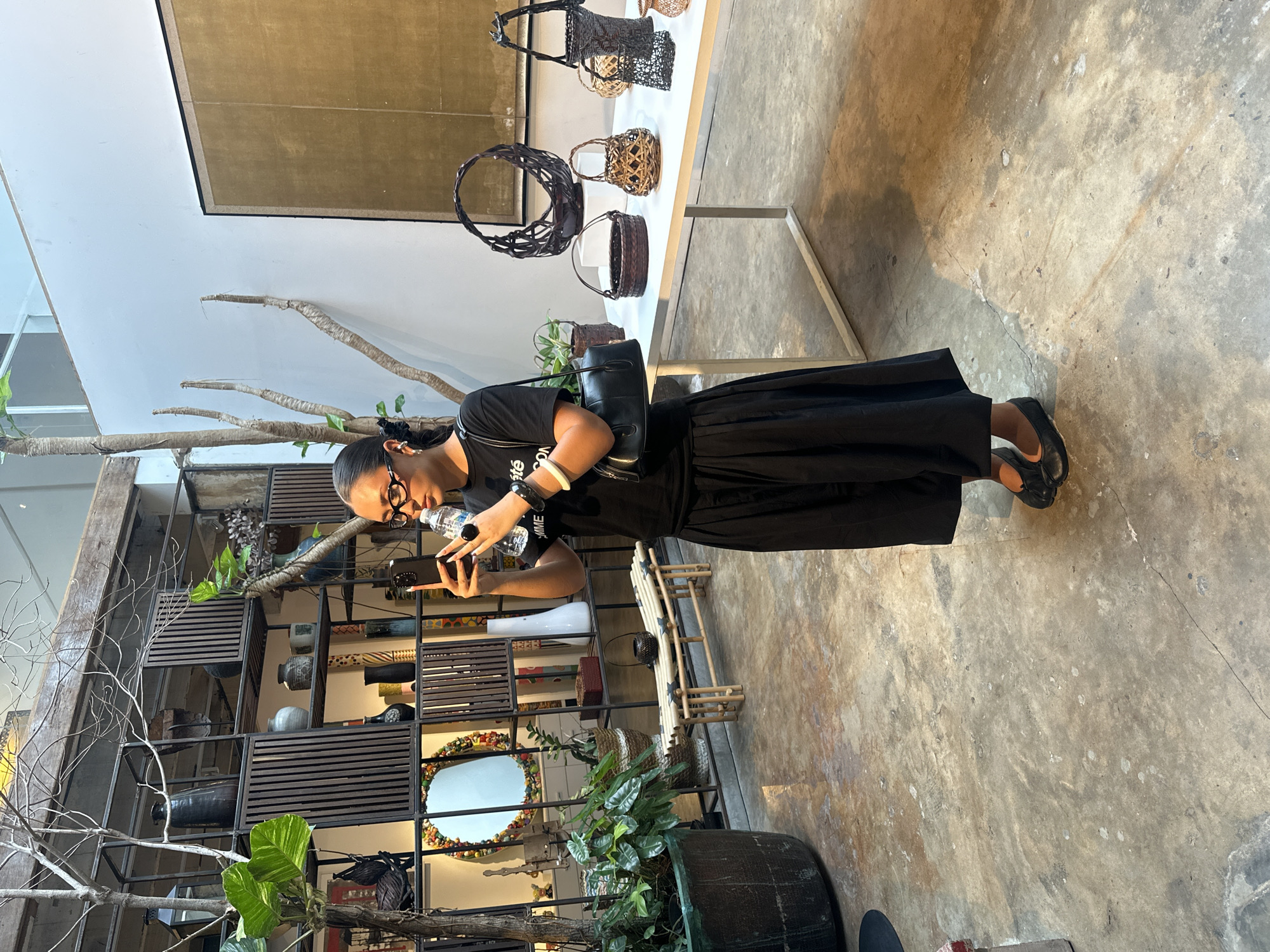 A medium, angled indoor shot captures a person in Bangkok, Thailand, engaged in photography. The individual, clad in black, is focused on their phone, likely taking a picture. They stand near a white table displaying woven baskets. A tall, decorative shelf filled with various objects and plants is visible in the background. The floor is a mottled concrete.