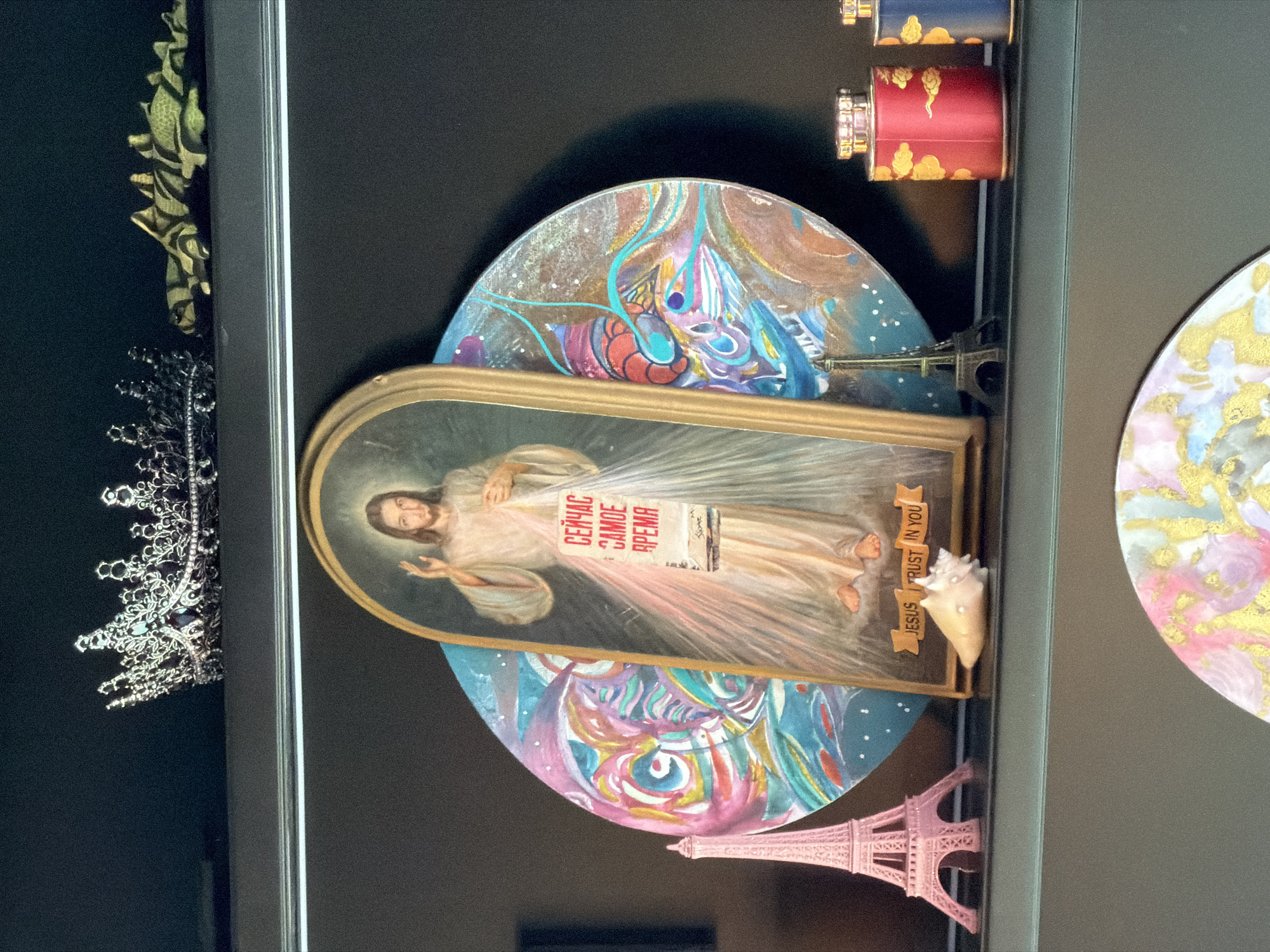 A framed artwork, likely hanging on a wall, features a portrait of Jesus Christ with rays emanating from his heart, overlaid with a newsprint-style text in Cyrillic script ("СЕЙЧАС САМОЕ ВРЕМЯ"). The portrait is framed in gold and set against a backdrop of painted semi-circles with abstract, colorful designs. Miniature Eiffel Tower replicas, one silver and one pink, are positioned at the bottom of each semi-circle. A small conch shell is affixed to the frame near the portrait of Jesus. To the left of the framed artwork, partially visible, are a silver crown and a green and gold decorative item. Adjacent to the right is a decorative circle with gold accents. Two stacked, decorative cylinders (red and blue) are situated at the upper right. The entire scene is contained within a dark-colored frame. The specific location in Moscow, Russia cannot be determined from this image.