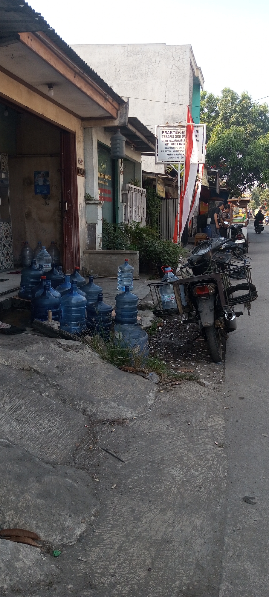 The image shows a street scene in front of a building, likely in Setu, Indonesia. Several large blue plastic water jugs are lined up on the ground near the building's entrance. Some of these jugs appear to be full, while others are empty. A motorcycle with a metal rack on the back is parked to the right of the water jugs. The motorcycle has a license plate that reads "AB 4670 VK". In the background, there is a building with a sign that says "PRAKTEK MA". Red and white Indonesian flags are hanging from the building. Two people are visible near a food stall to the right of the building, and another person is riding a motorcycle in the distance. The ground in the foreground is paved with concrete and shows signs of wear and tear, with scattered debris and cigarette butts. Greenery is visible behind the building. The lighting suggests it is daytime, possibly late afternoon, with shadows cast by the objects.