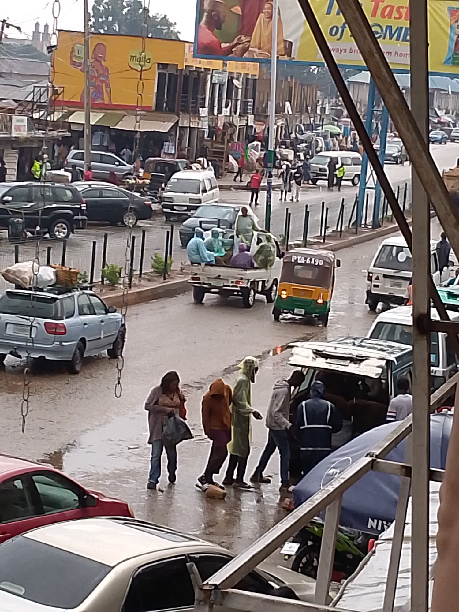 The image presents a vibrant street scene in Bukuru, Nigeria, on a wet day, suggesting recent rainfall or an ongoing drizzle, under an overcast daytime sky. The asphalt roads and sidewalks are visibly wet, reflecting light and showing puddles.
The scene is bustling with both vehicular and pedestrian traffic. Numerous people are walking along the street and sidewalks, some carrying bags. Notable individuals include a woman in the foreground holding a bag, and a person further back wearing a bright yellow-green rain jacket. Towards the lower right, several people are gathered around an open vehicle, possibly a van or SUV, suggesting loading or unloading activity. A man in a blue uniform with reflective strips is also visible in this group.
Various vehicles populate the street: multiple sedans, SUVs, and vans are seen parked or in motion. A light blue station wagon with luggage and goods piled on its roof is prominent on the left. In the middle ground, a white pickup truck carries several passengers or goods in its bed. A distinct yellow, green, and brown auto-rickshaw, commonly known as a Keke Napep, is moving, displaying the license plate "PTA-6190" on its rear.
In the background, a row of commercial buildings and shops line the street. Large billboards are mounted above these structures; one bright yellow billboard features an illustration of a woman holding a product and the text "malla." Another partially visible sign above it reads "The Taste of HOME" and "Always brings you home." In the lower right corner, a blue umbrella or tent-like structure is visible with text that appears to start with "NIV".
The overall impression is one of a busy urban or market area, where daily commerce and commuting continue despite the wet weather.