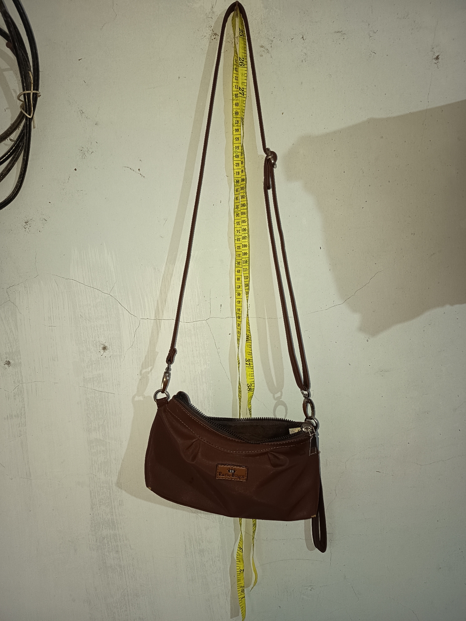 A brown purse hangs from a nail on a textured white wall. A yellow measuring tape is also hanging from the wall, draped over the purse's strap and extending down to the bottom of the frame. The purse has a zipper closure and a small brown tag with text on it. The wall has several cracks and discoloration. To the left of the purse, a black coiled cable is attached to the wall with a beige tie. The scene appears to be indoors, possibly a workshop or storage area, in Krampilan, Indonesia.