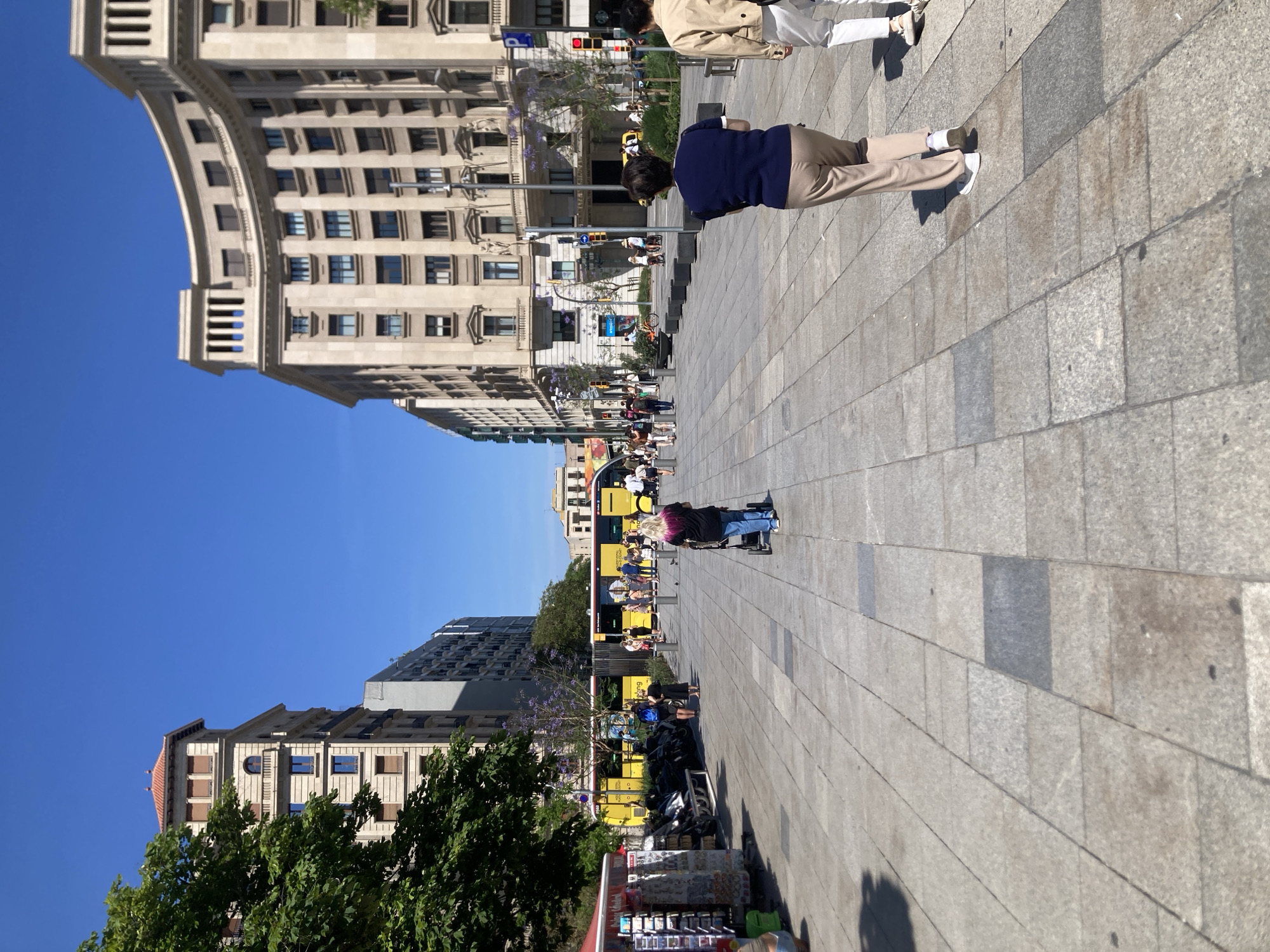 The image depicts a street scene in Barcelona, Spain. A wide, paved pedestrian walkway stretches through the middle of the frame, with buildings lining either side. A yellow tour bus is visible in the distance, crowded with people. Pedestrians are scattered along the walkway; one person is in a wheelchair. To the upper right, two people walk away from the camera. The sky is clear and blue. Vendors and merchandise stands are visible on the lower-left side of the walkway. Trees with purple blossoms are interspersed among the buildings. The architecture of the buildings varies, with some having ornate facades and curved elements.